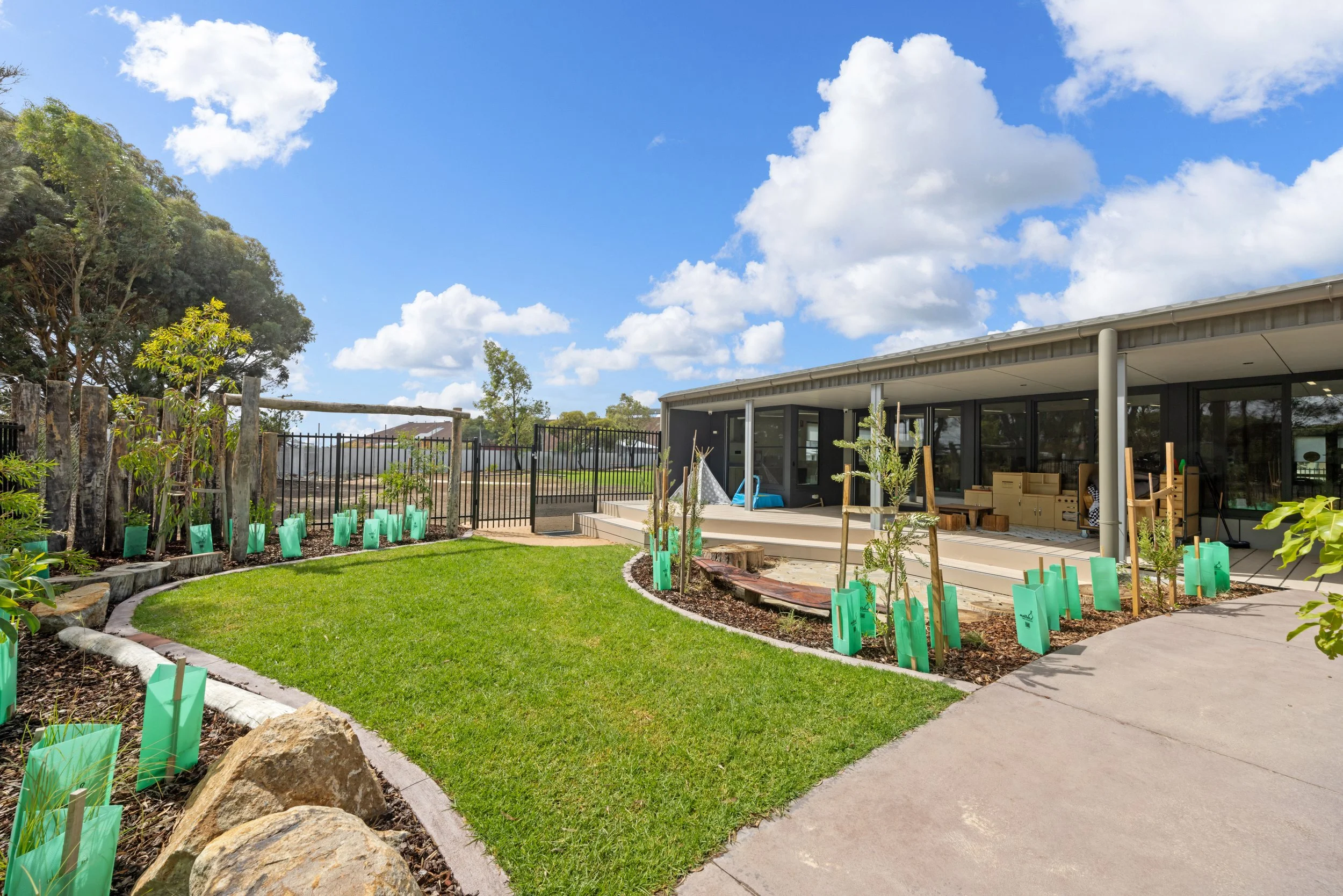 Modern house with a backyard garden, green grass, young trees protected with green plant covers, and a patio with outdoor furniture, under a partly cloudy sky.