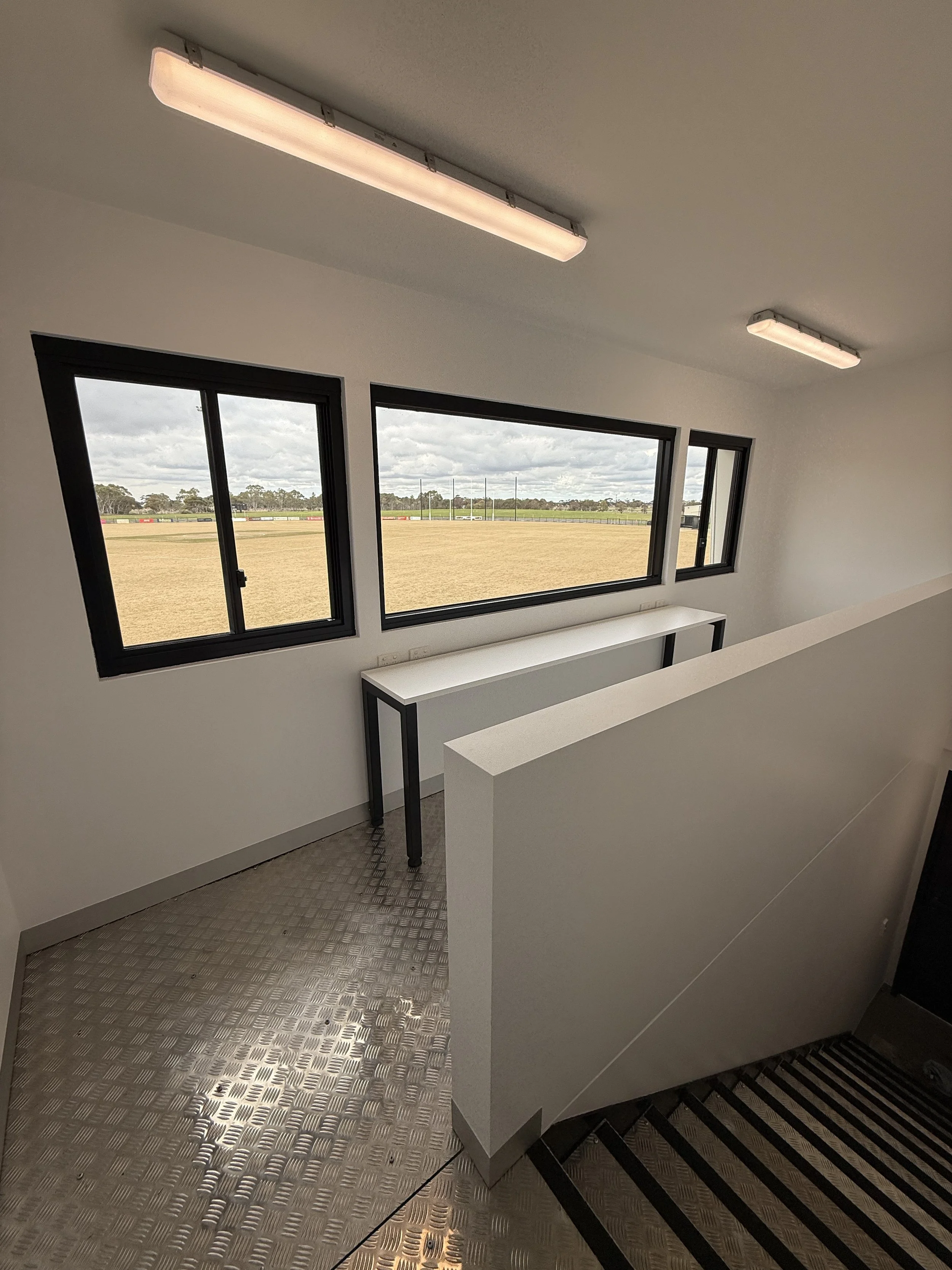 Interior of a room with three windows, two are small and one is large, showing an outdoor field with grass and a cloudy sky. There are fluorescent ceiling lights, a table with black metal legs, a partial plain white wall, and a black and grey striped carpeted staircase.