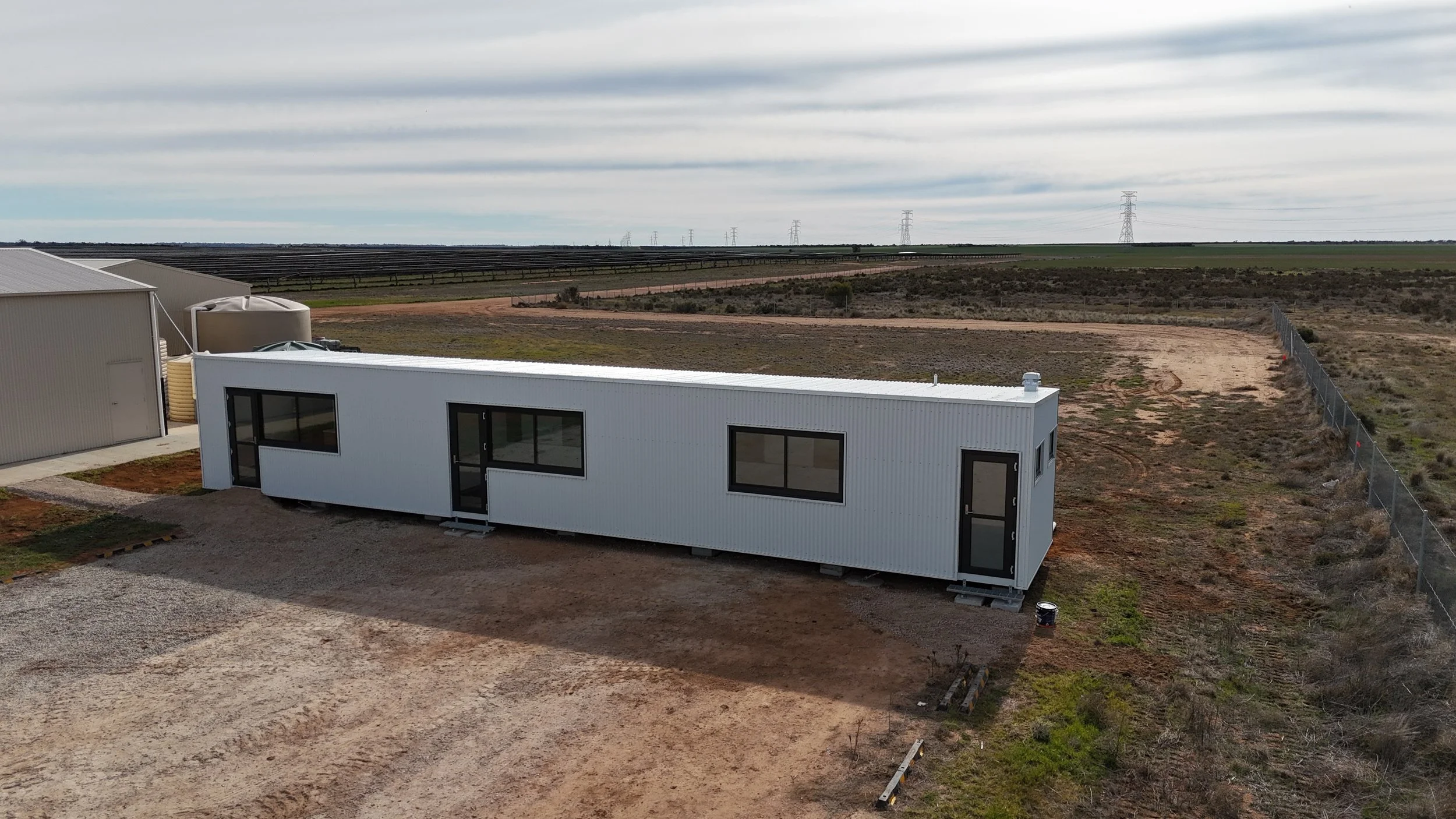 A white modern tiny house on a gravel lot in a rural area with open fields, power lines, and cloudy sky in the background.