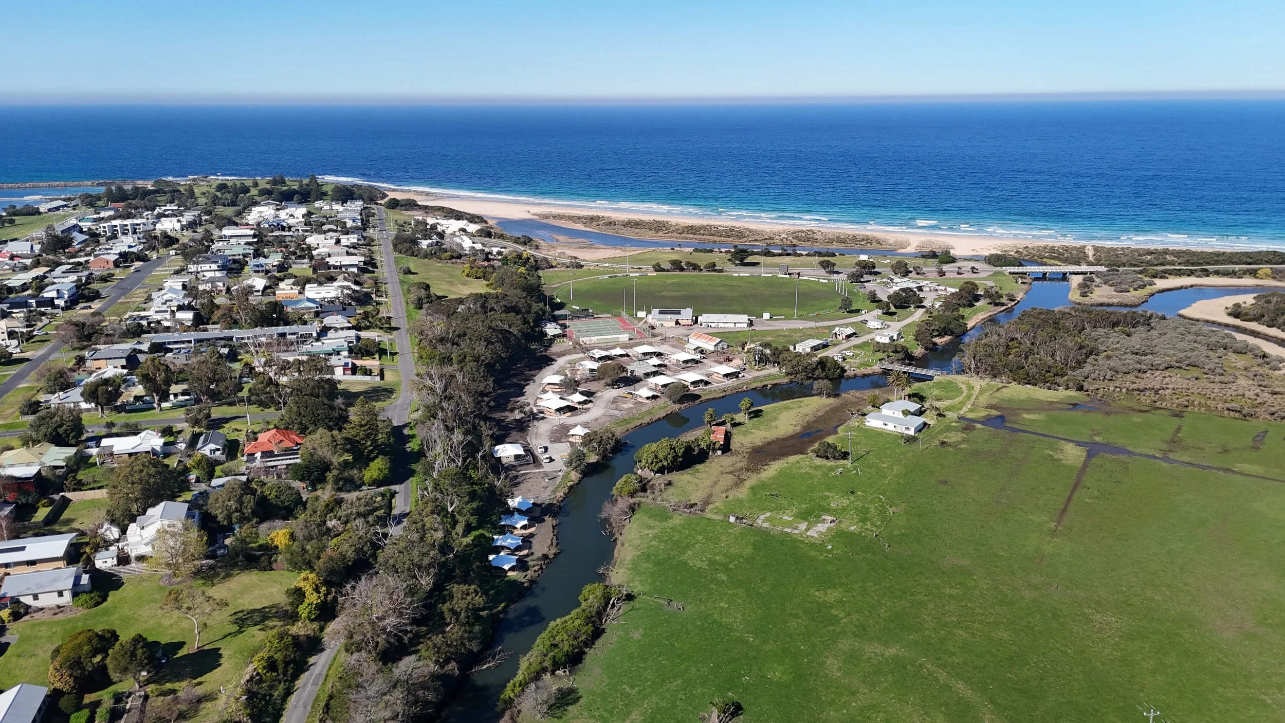 Aerial view of a coastal town with houses, roads, green parks, a river, and a beach with the ocean in the background.