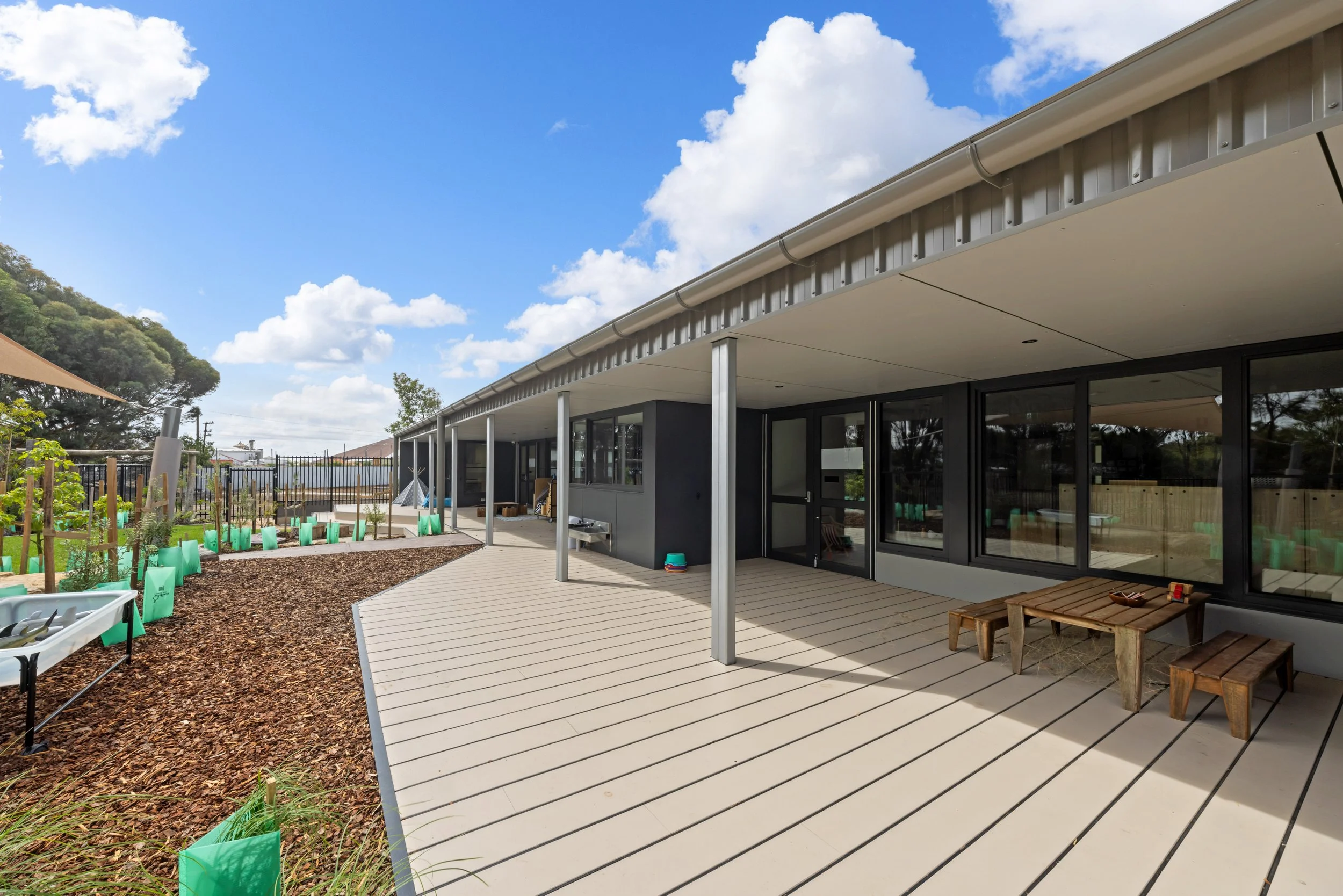 Modern building with large glass windows and sliding doors, featuring a spacious outdoor wooden deck, with a garden area on the left side with small plants and mulch, under a partly cloudy blue sky.