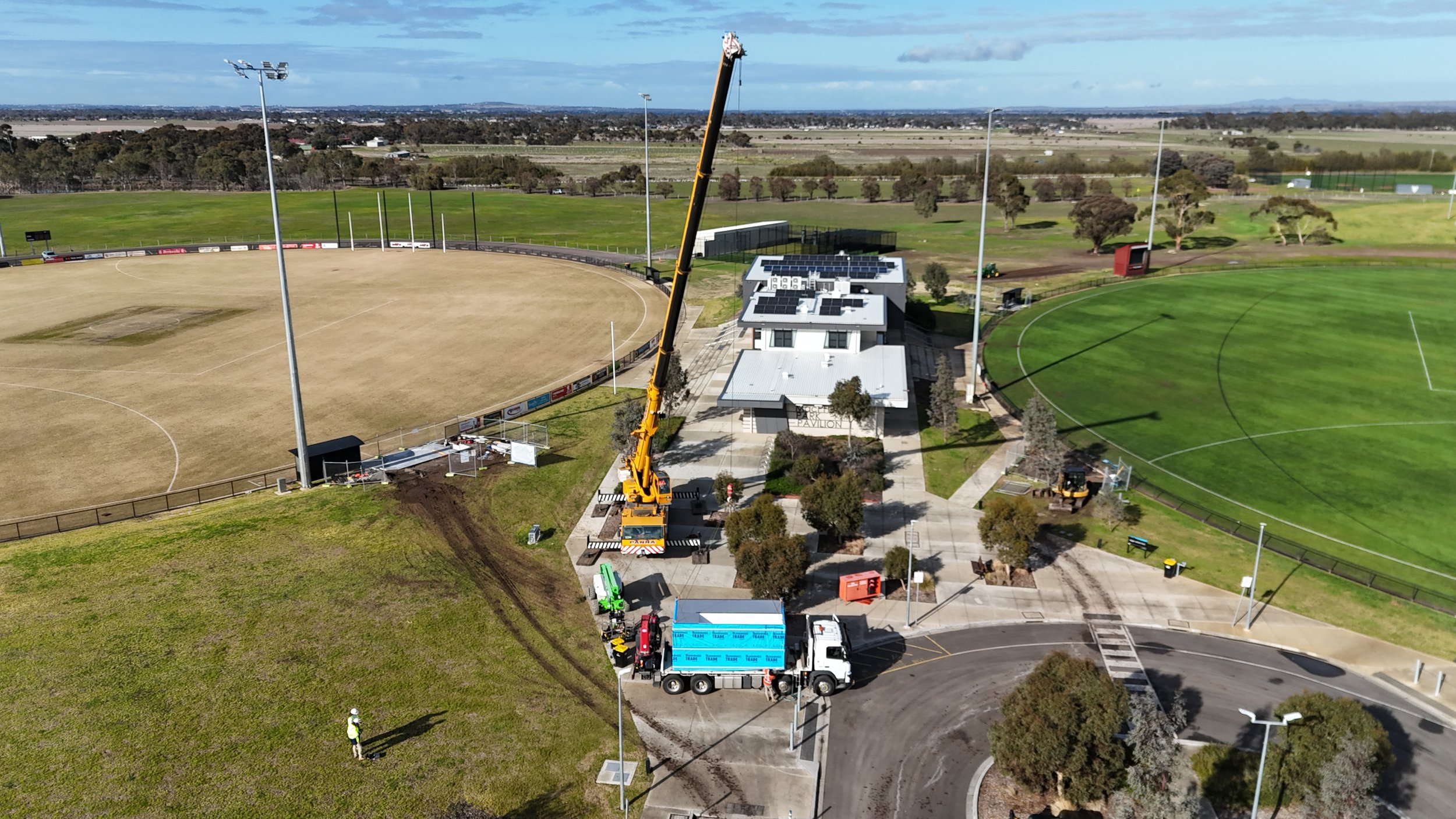 An aerial view of a sports stadium with construction activity occurring near the center. There is a large crane lifting materials, construction workers on site, and construction trucks. The stadium features a grass soccer field on the right and a dirt field on the left, with surrounding pathways, trees, and lampposts, and open fields beyond the stadium.