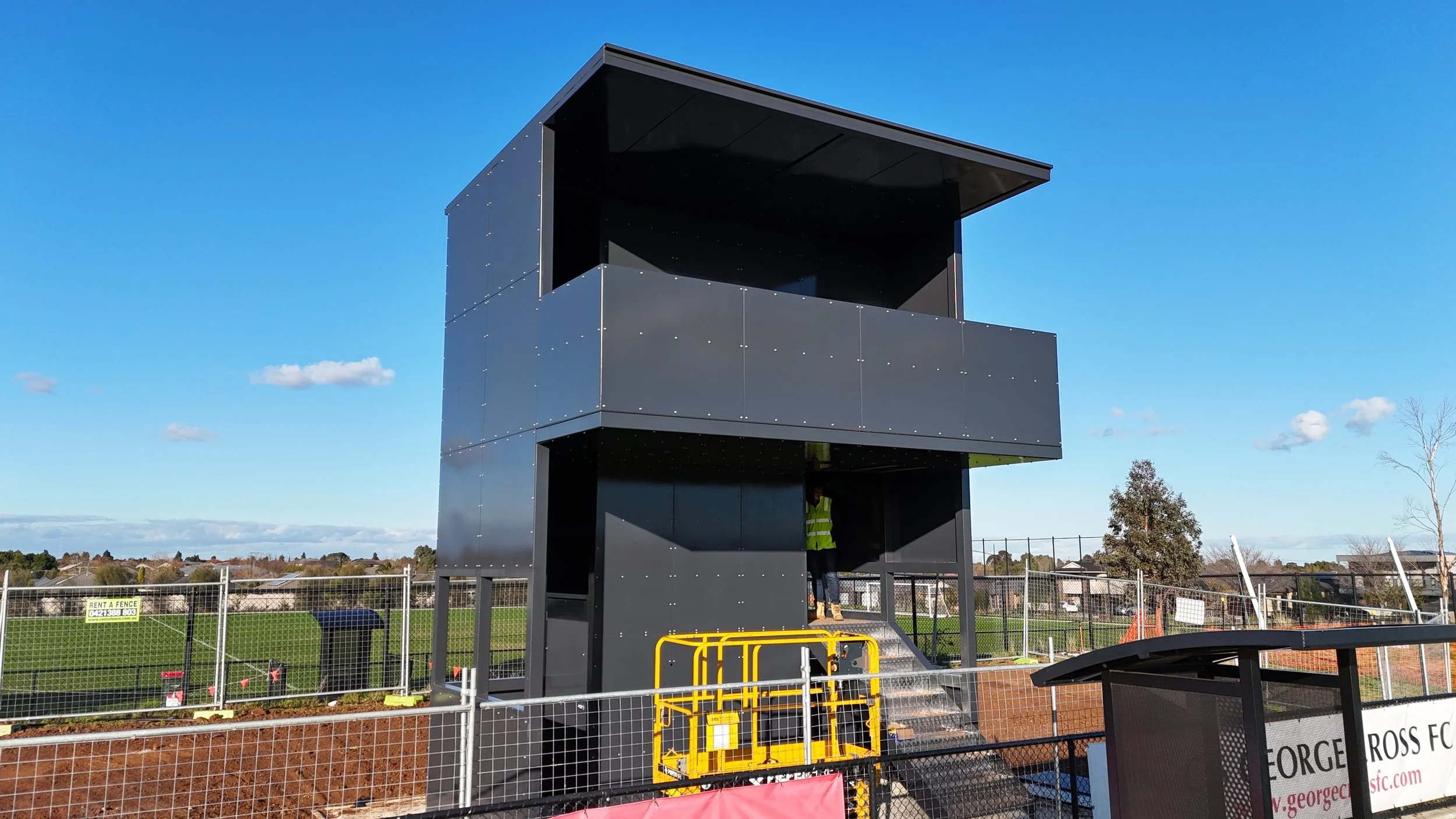A black, modern, multi-level watchtower structure with an overhanging roof, situated outdoors in a fenced area with a blue sky and some trees in the background.