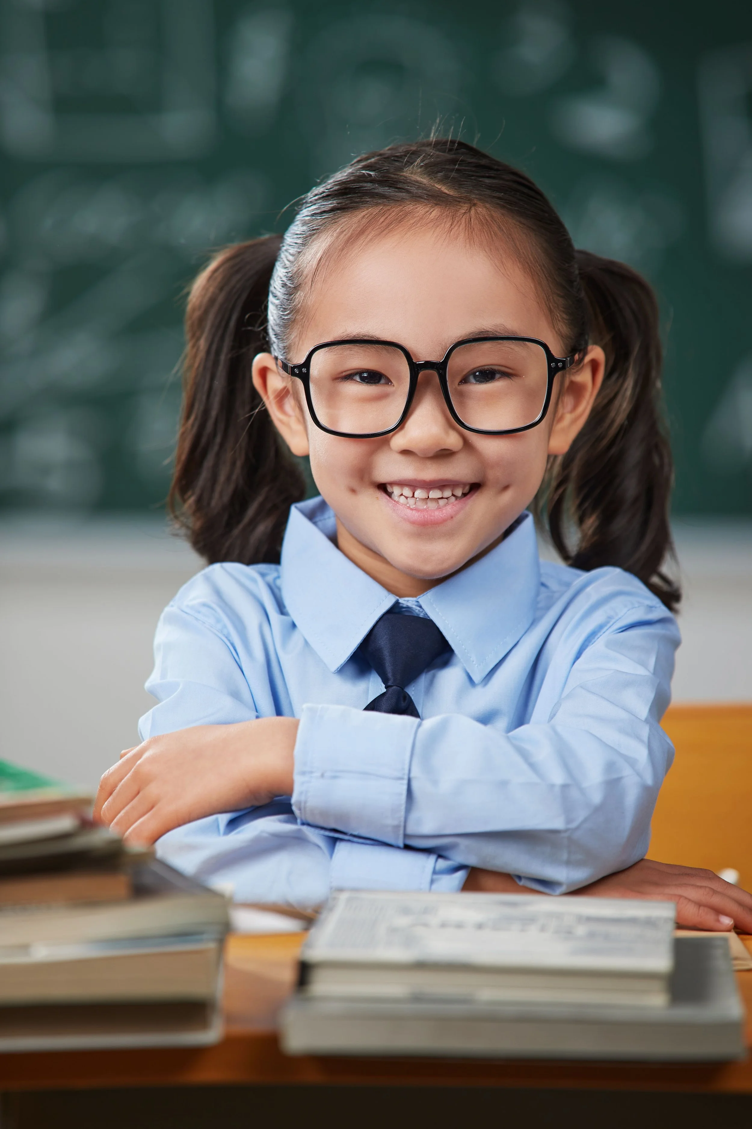 A young girl with glasses smiling and sitting at a desk with books in the foreground, in a classroom setting.