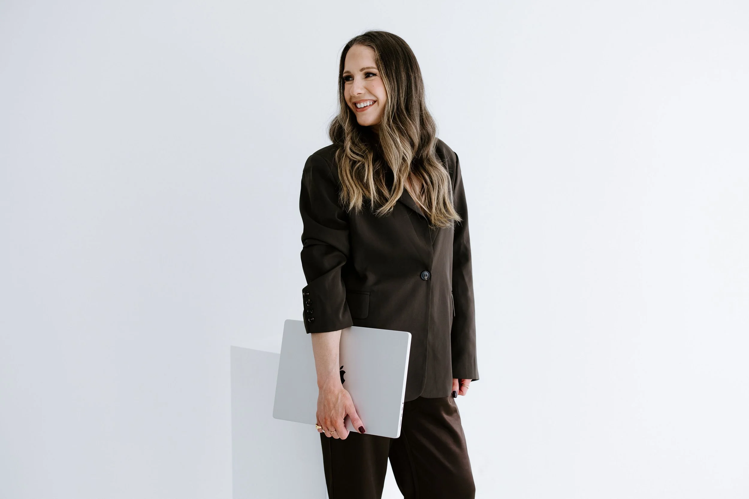 A woman in a black suit holding a silver laptop and smiling, standing against a plain white background.