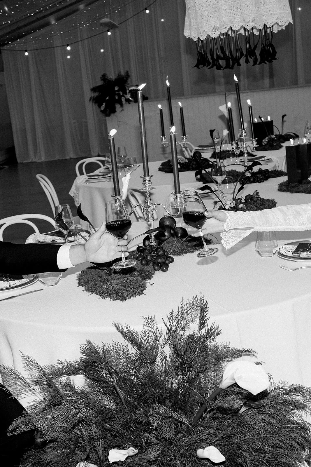 A black and white photo of a decorated dinner table with candles, wine glasses, and floral centerpieces. Two people are toasting with glasses of red wine.