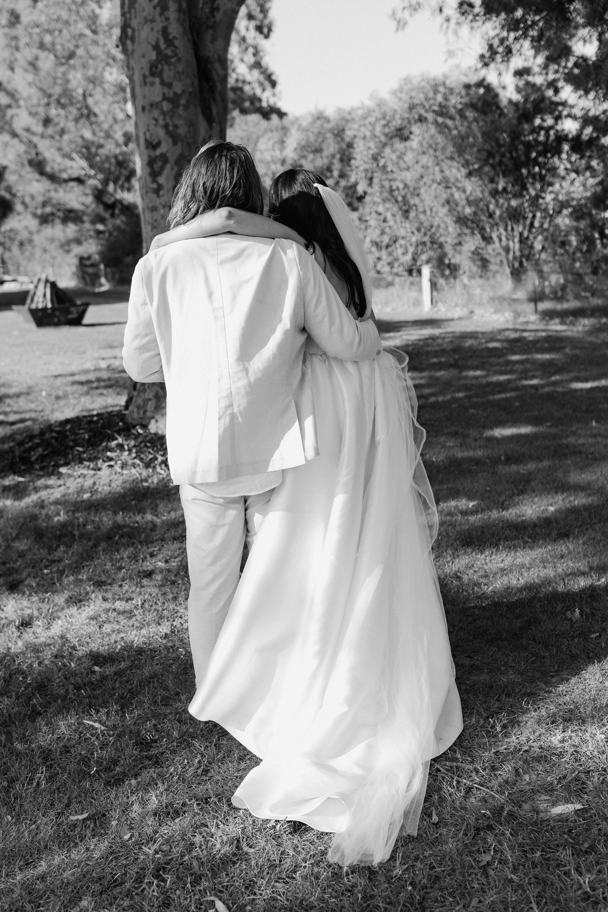 A black and white photo of a couple embracing outdoors, with the woman wearing a wedding dress and veil, and the man wearing a suit, near a large tree and a grassy area.