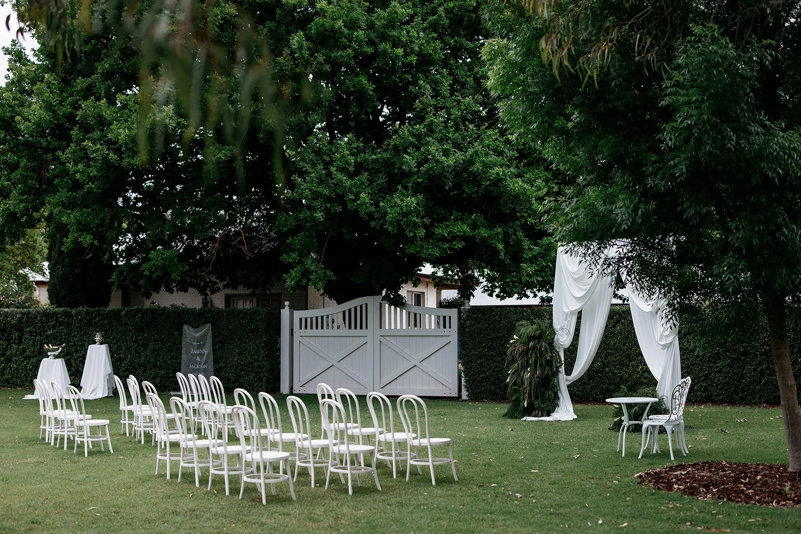 Outdoor wedding ceremony setup with white chairs arranged in rows on green grass, a white arch with draped fabric, and tables with decorative items under trees and a fence.