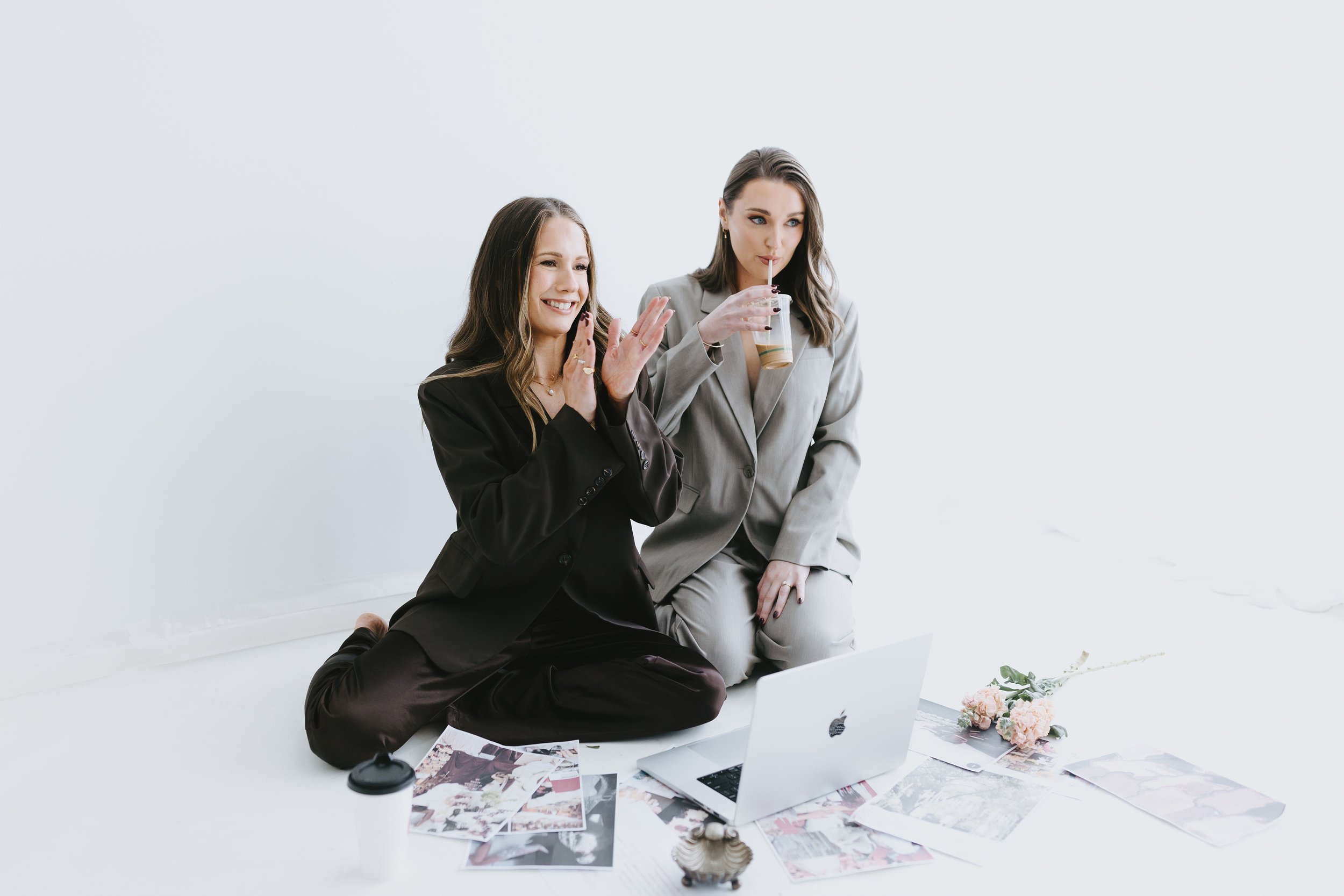 Two women in business attire sitting on the floor surrounded by laptops, magazines, and flowers, appearing to be collaborating or brainstorming.