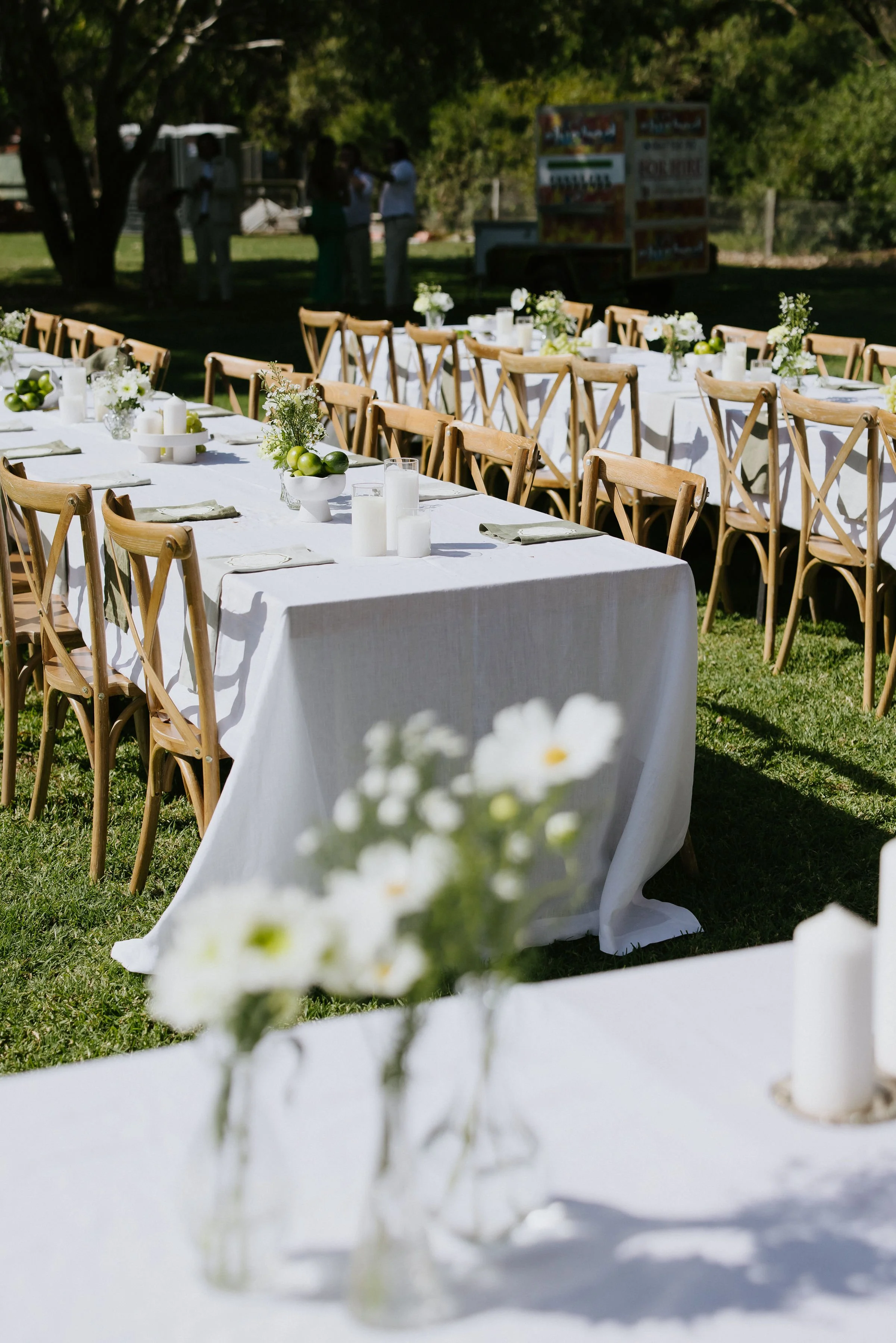 Outdoor banquet table decorated with white tablecloth, floral centerpieces, and green apples, set for a celebration in a sunny garden with trees and a food truck in the background.