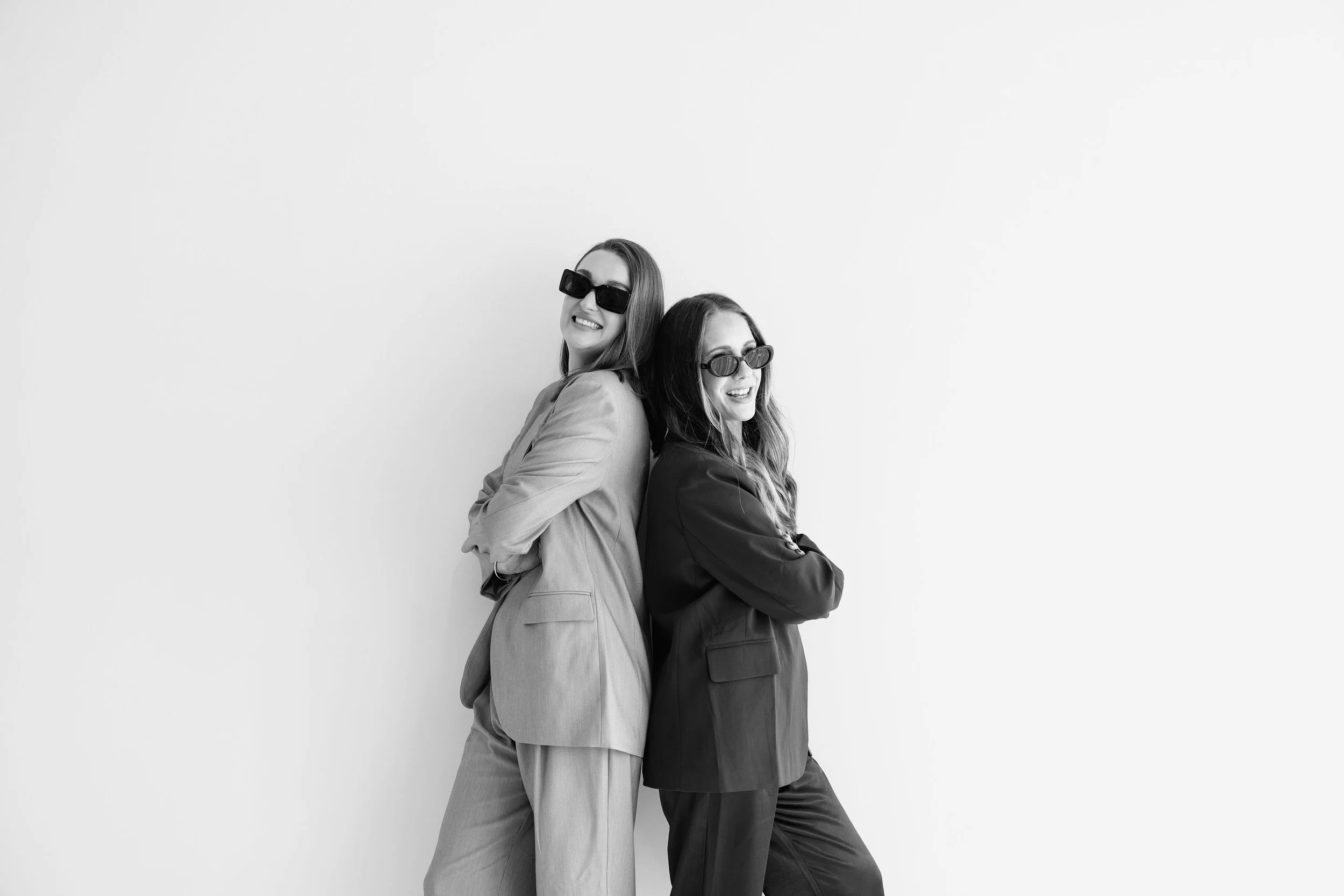 Two women stand back to back, wearing sunglasses and smiling, one dressed in a light-colored suit and the other in a dark suit, against a plain white background.