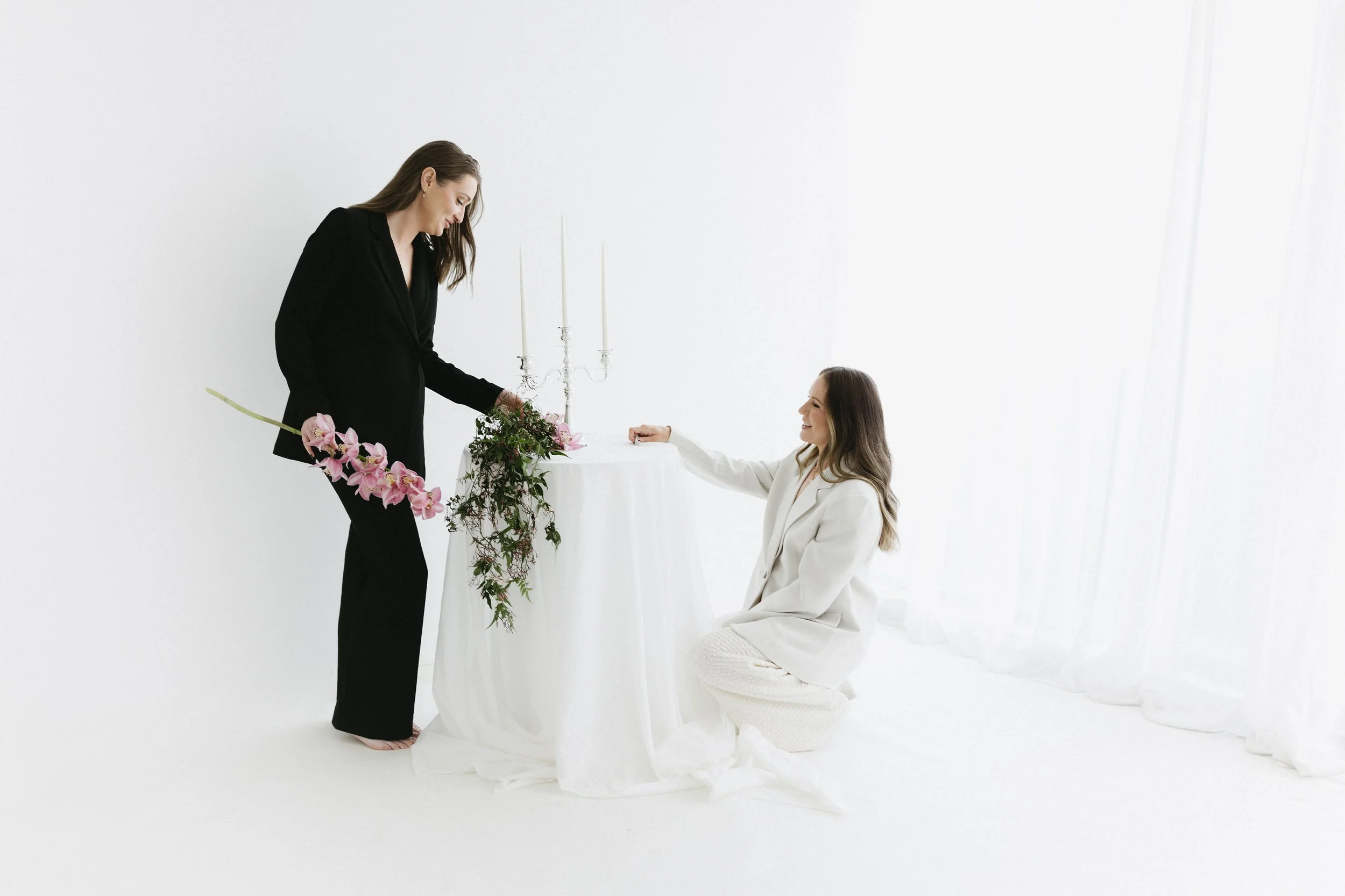 A woman in a white suit kneeling on a white cloth-covered surface, smiling and holding a woman's hand, with another woman standing nearby in a black outfit holding flowers and greenery, in a minimalist white room.