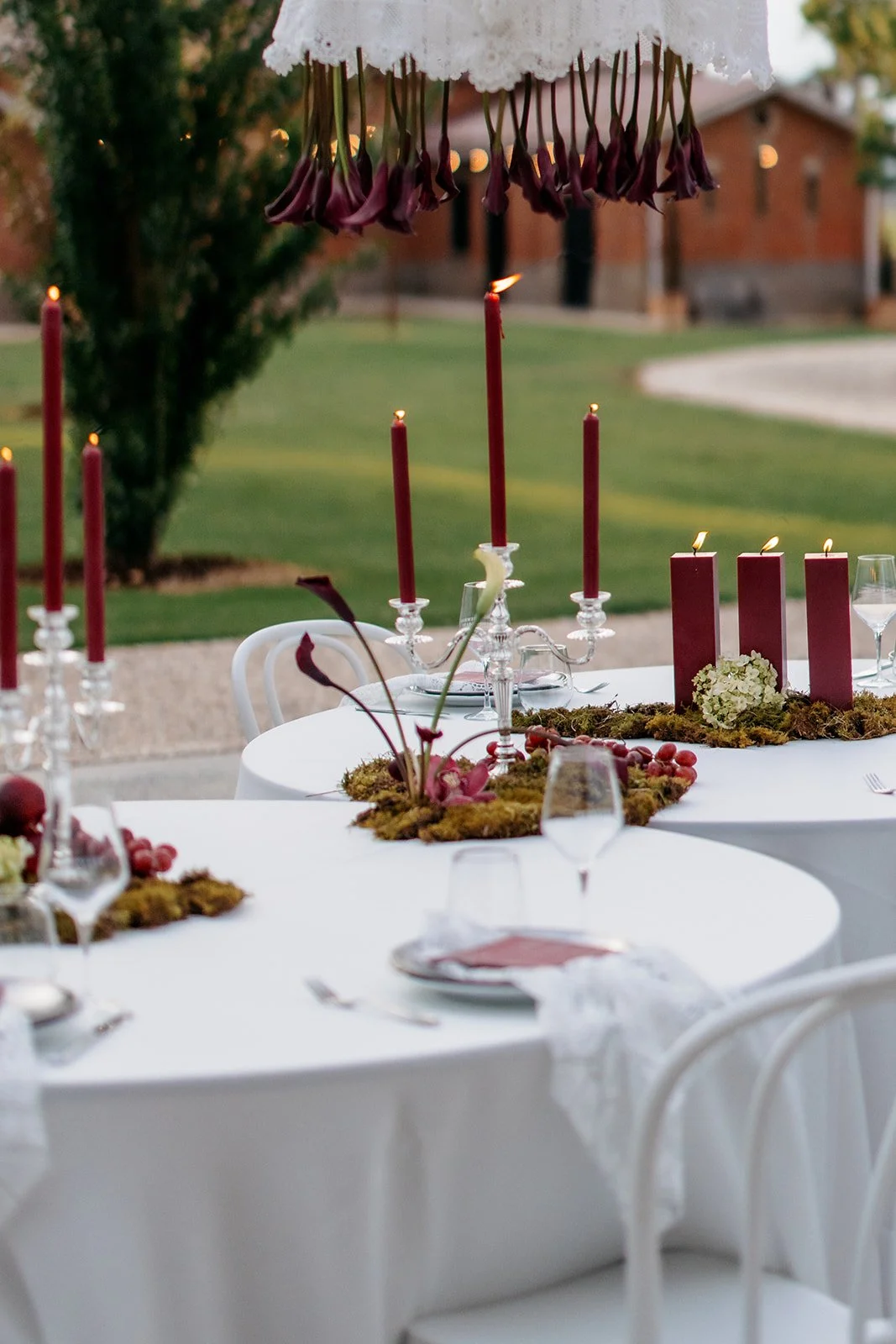 Outdoor dining table decorated with red candles, white flowers, moss, and grapes, under a lace canopy with hanging purple calla lilies, set for a formal event during the evening in a garden with a brick house in the background.