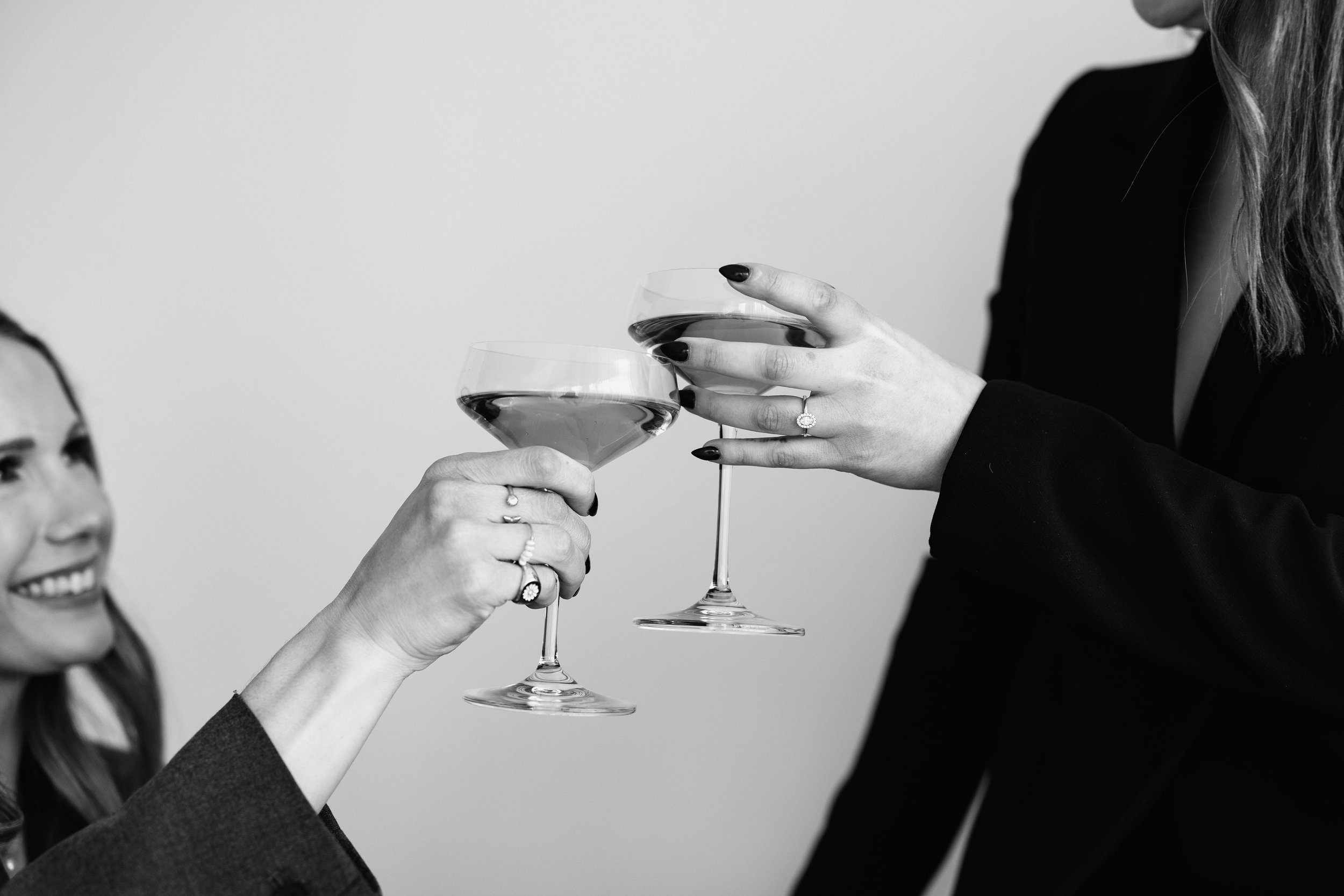 Two women clinking wine glasses in a toast, near a face of a smiling woman on the left, dress and rings visible, black-and-white photo.