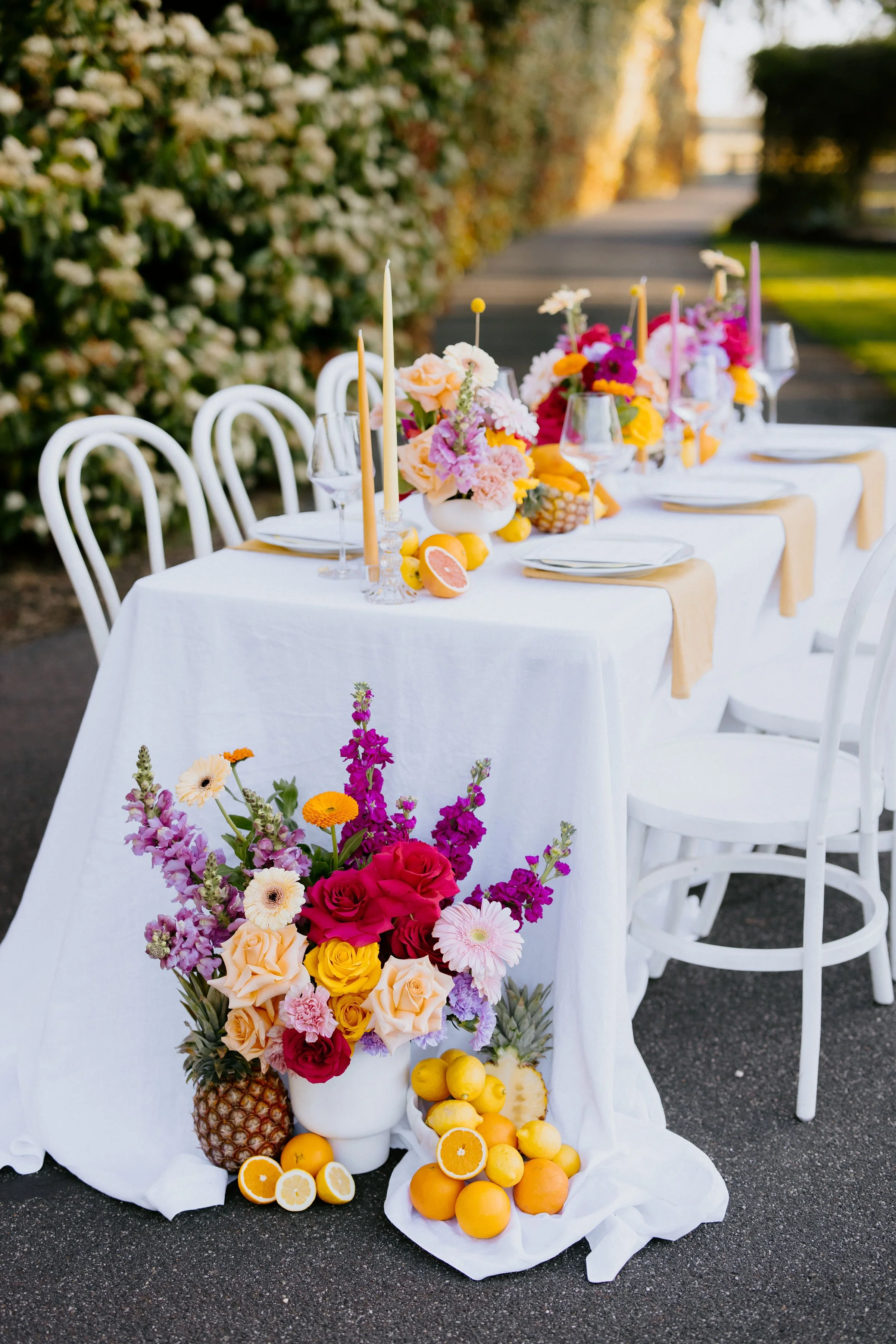A table decorated with colorful flowers, candles, and fruits outdoors on a sunny day, with chairs around it and a hedge in the background.