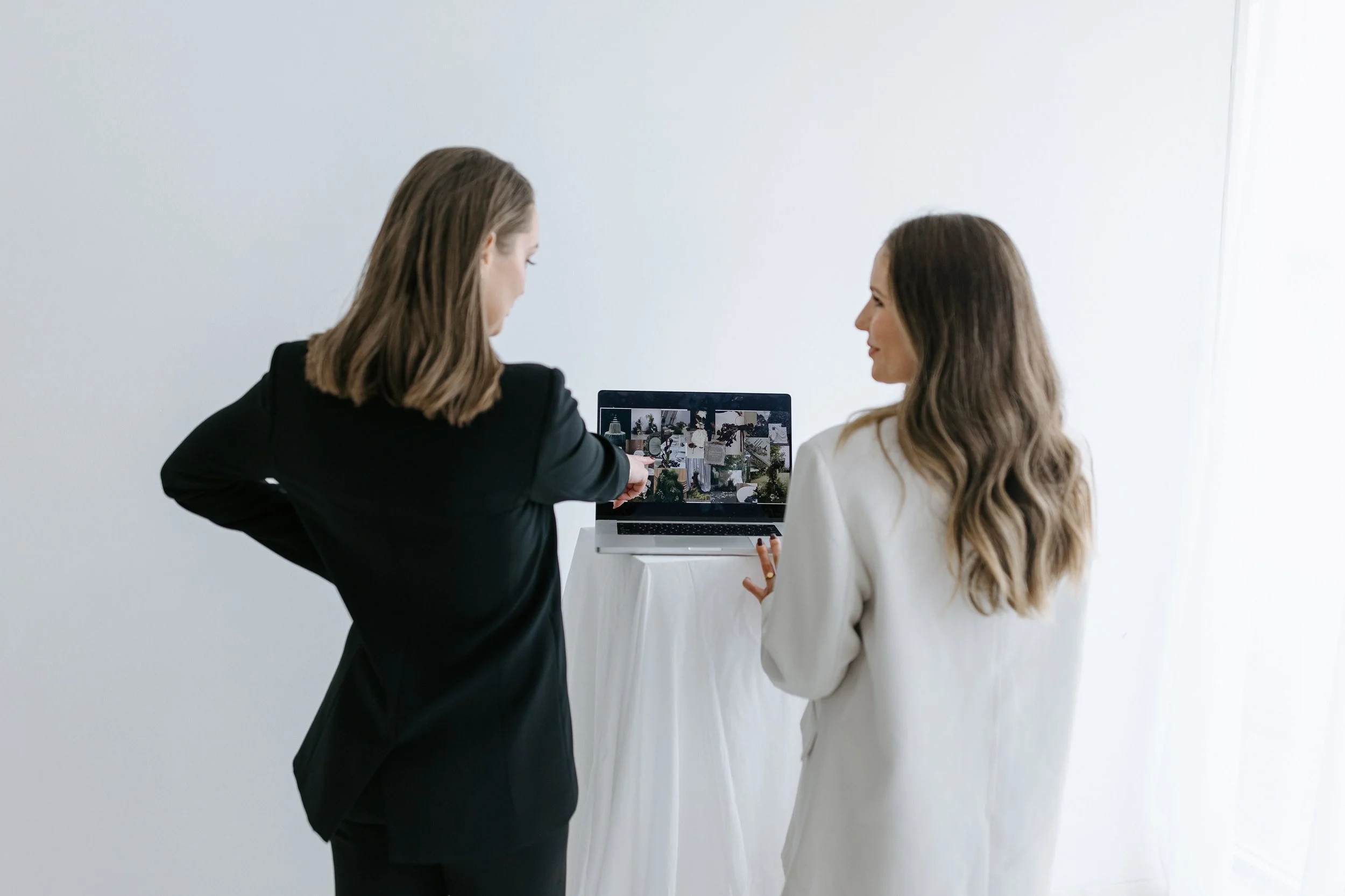 Two women with long hair, one in a black blazer and the other in a white blazer, are looking at a laptop displaying a collage of images. The woman in black points at the screen while the woman in white smiles. They are in a minimalist room with plain