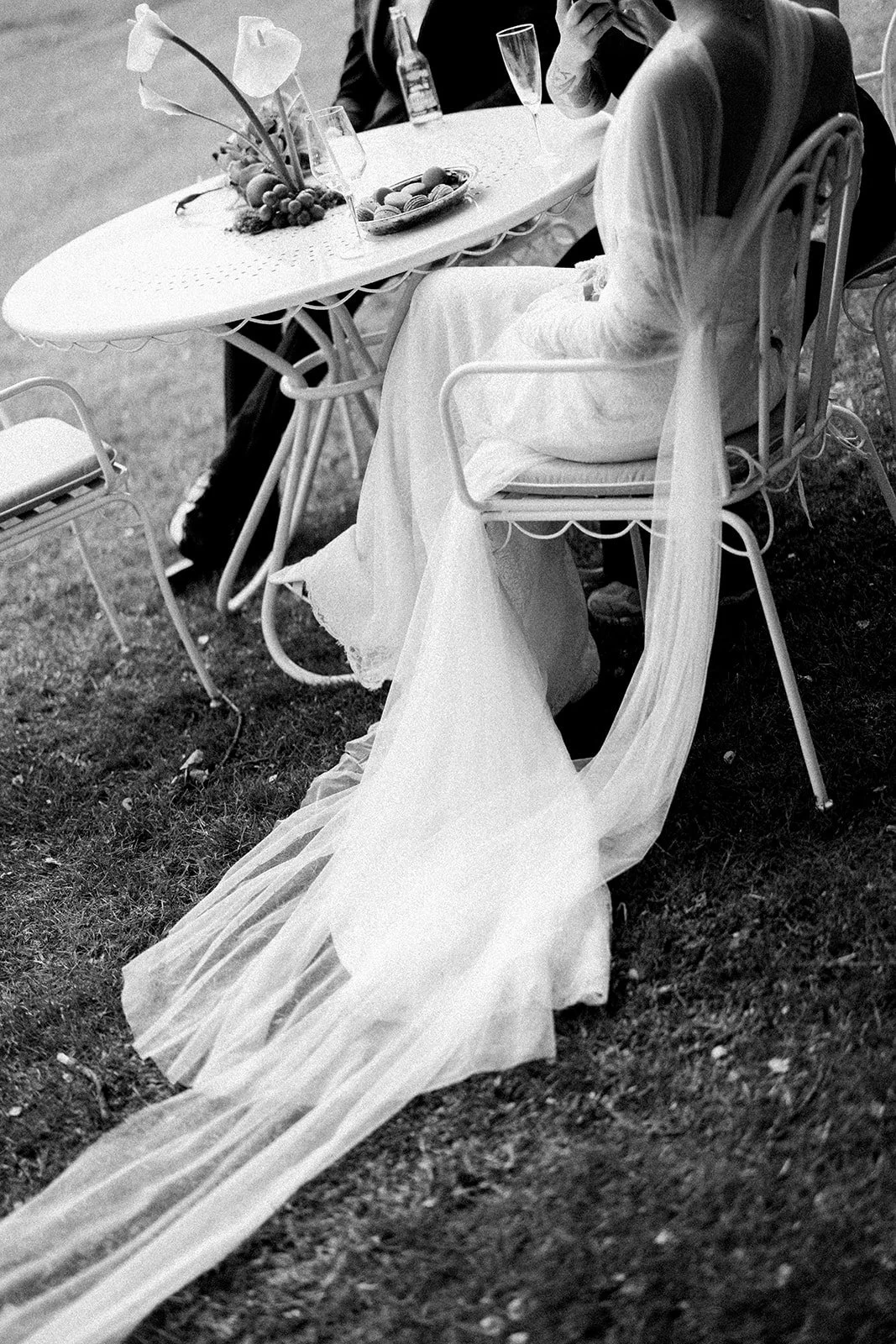 A wedding reception table set outside with a bride seated, with a long veil trailing on the ground. The table has flowers and drinks, including champagne glasses and a bottle.