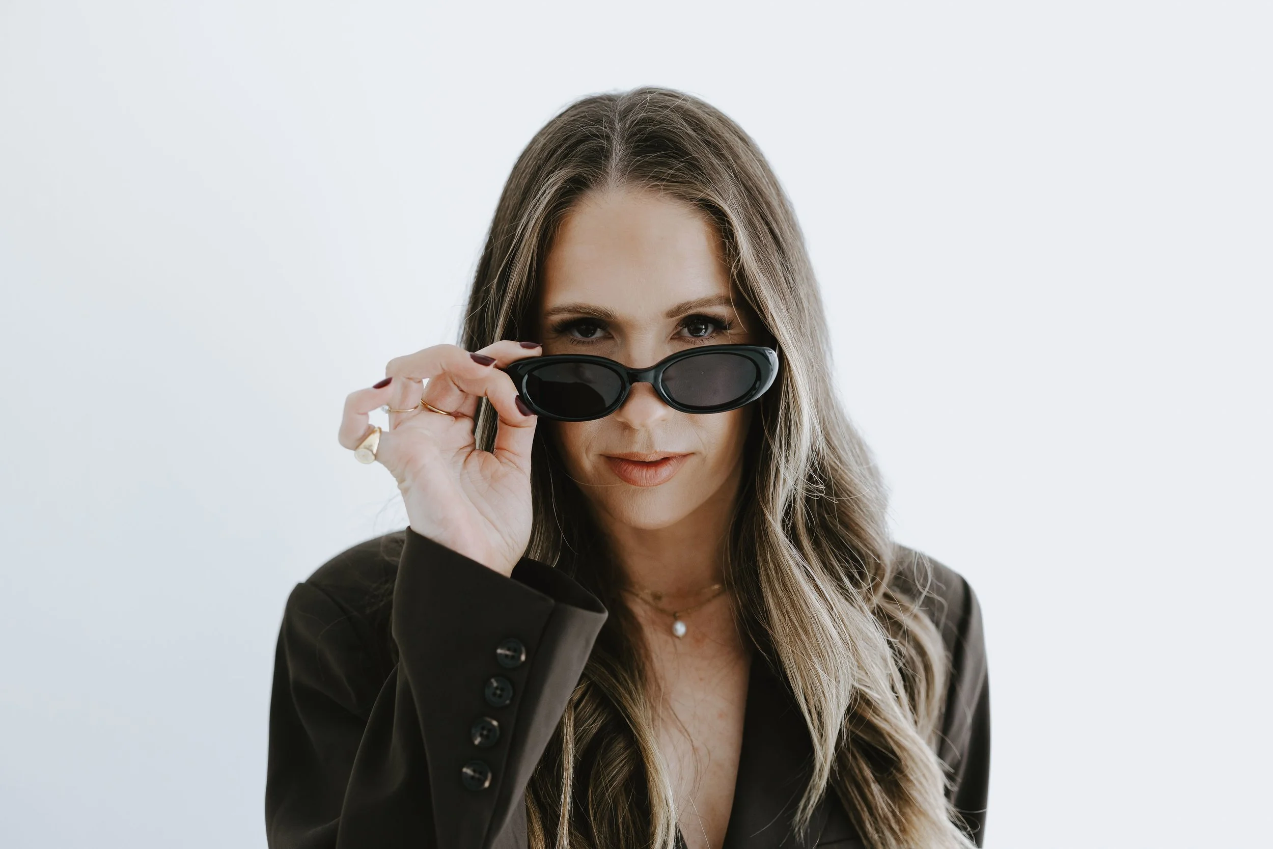 A woman with long, wavy hair and fair skin looking over black sunglasses she is holding with her right hand, wearing a dark blazer on a light gray background.