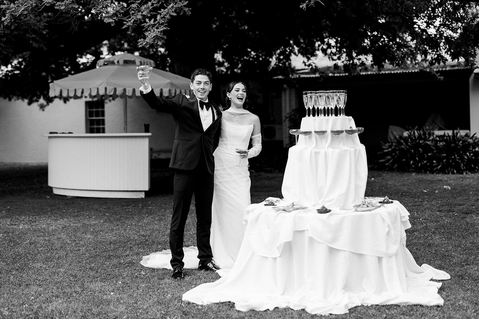 Black and white photo of a newlywed couple celebrating outdoors at their wedding reception, standing next to a champagne tower and cake table, with trees and a house in the background.