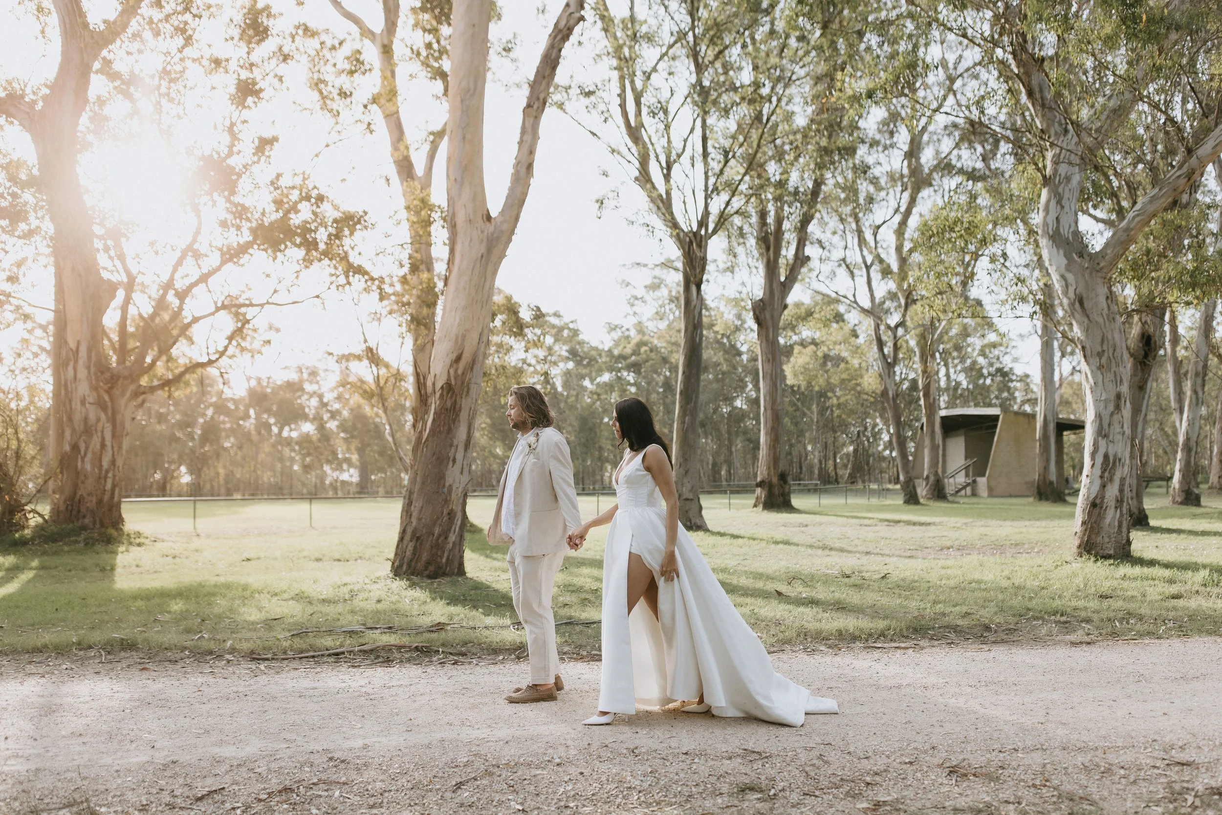 A couple dressed in wedding attire holding hands and walking outdoors among tall trees during sunset.