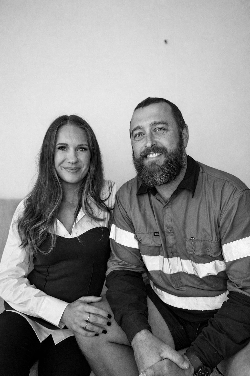 A black-and-white photo of a smiling woman with long hair and a man with a beard, sitting closely together against a plain wall.