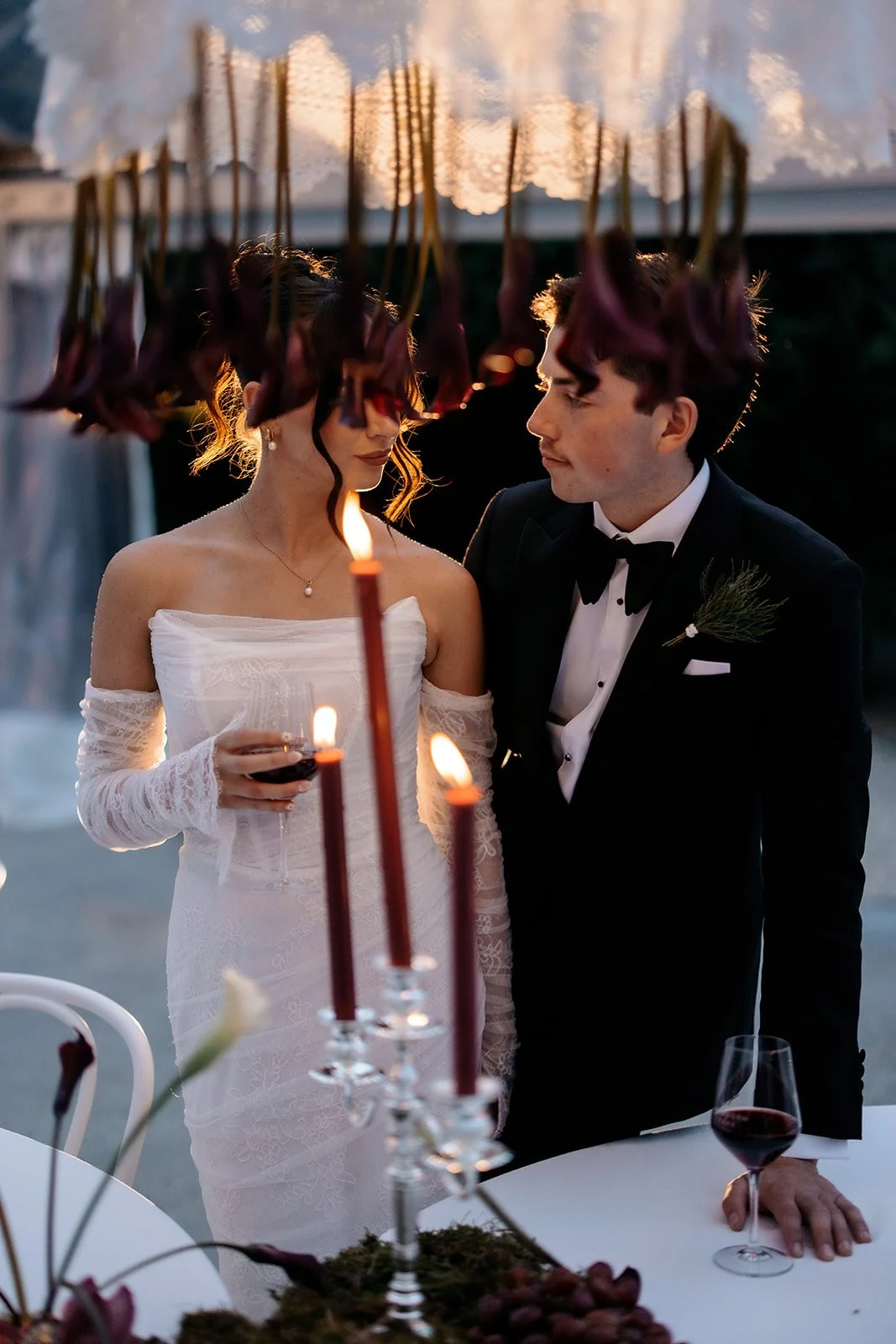 A bride and groom dressed in wedding attire stand close together during a candlelit outdoor celebration, with a table featuring red candles and glassware in the foreground.