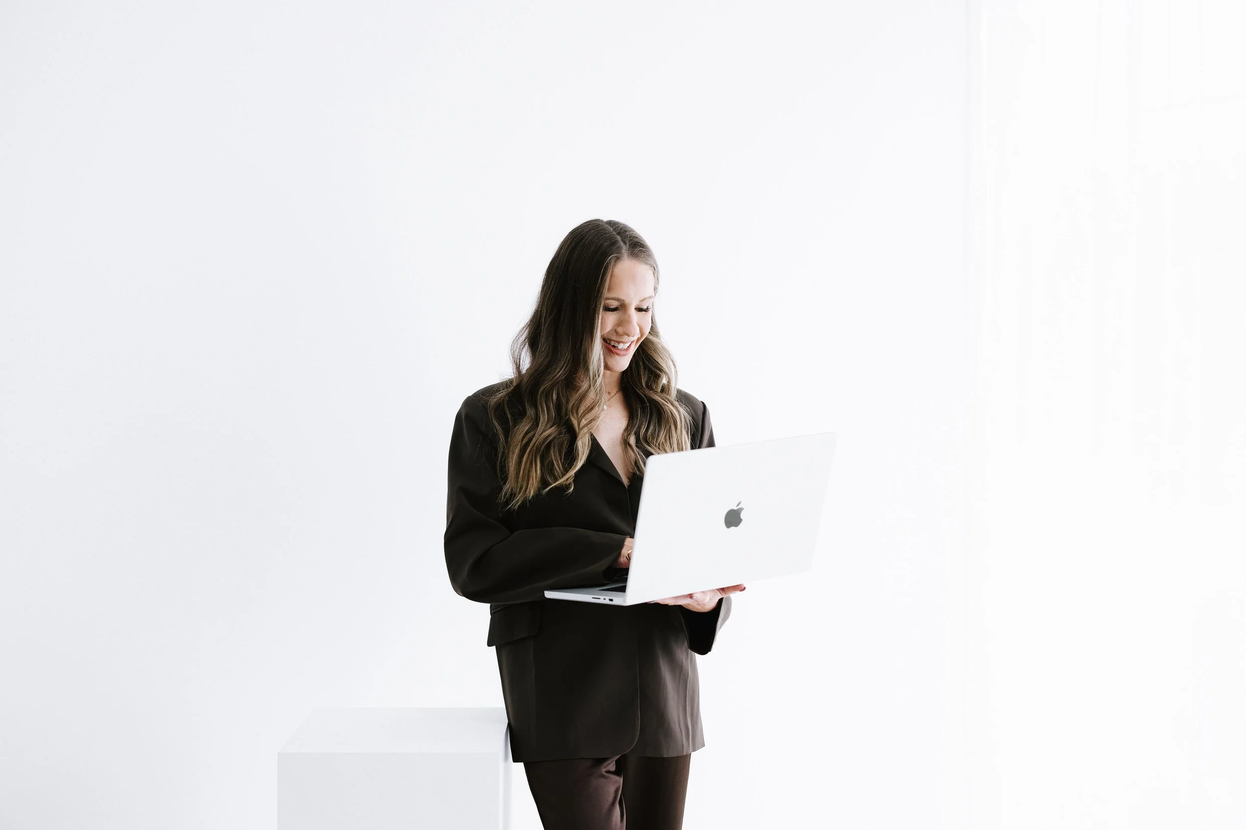 A woman with long wavy hair wearing a black blazer and black pants, standing against a white background, holding a silver laptop with an Apple logo, smiling while looking at the screen.