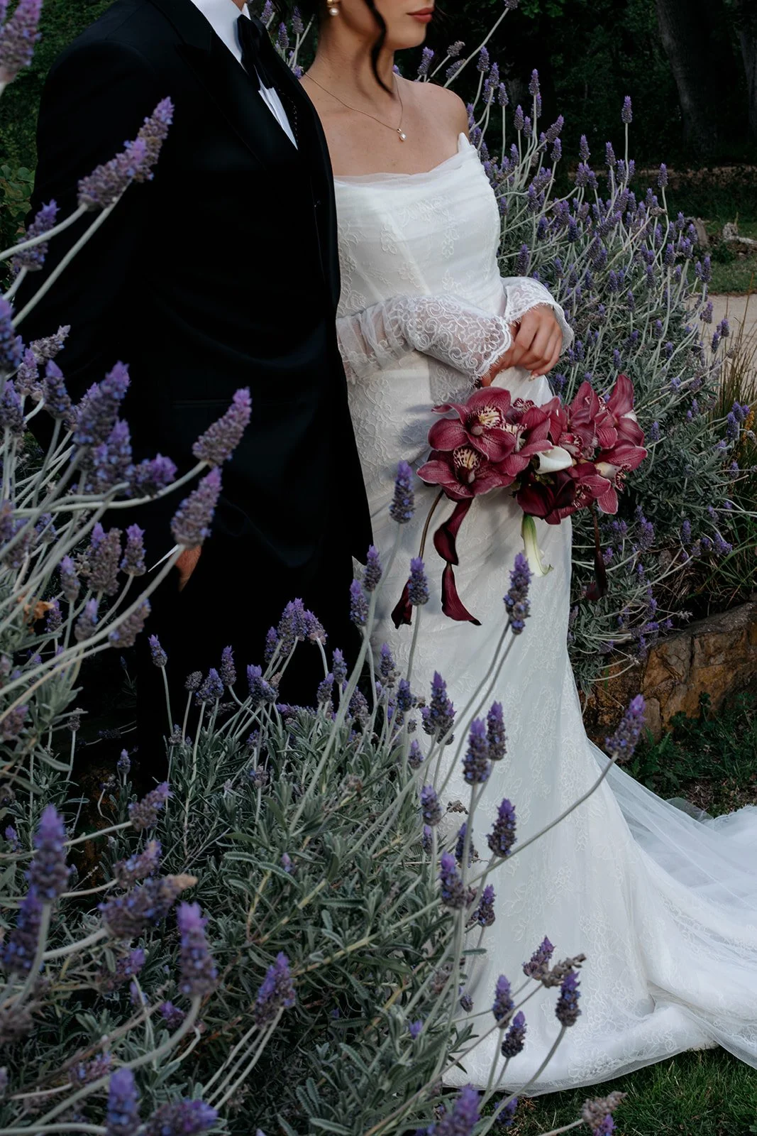 Partially visible bride and groom standing among purple lavender flowers, with the bride holding a bouquet of pink and white orchids.