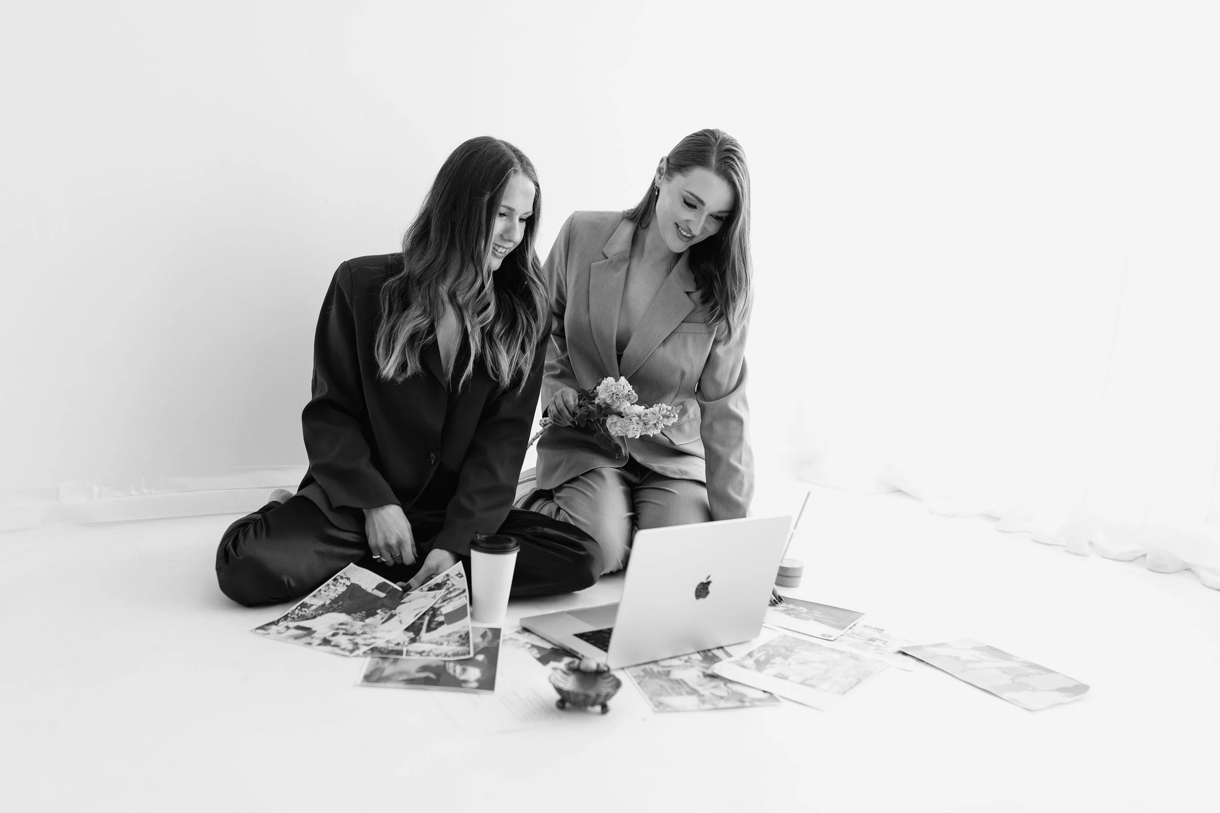 Two women sitting on the floor, looking at a laptop, surrounded by magazines, coffee cups, and papers, with a plain white wall in the background.