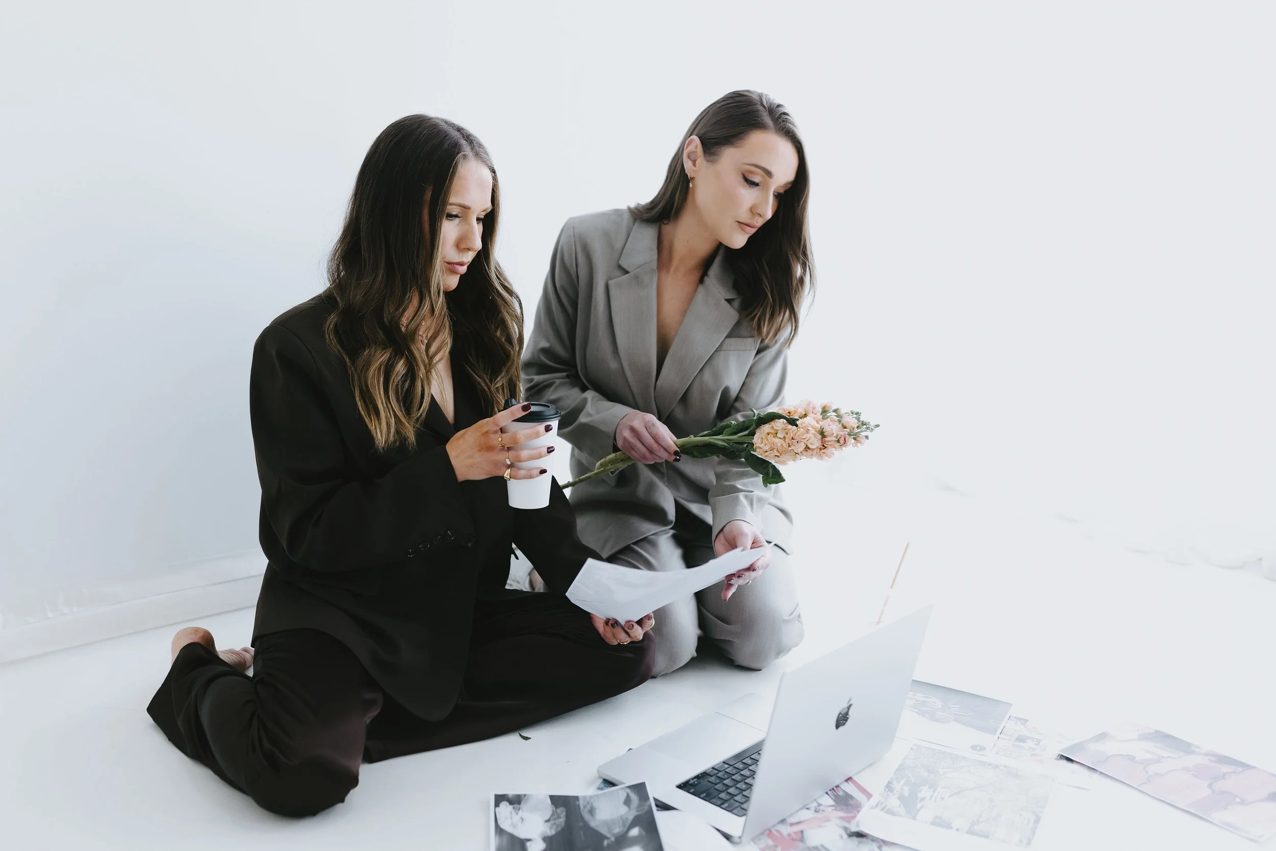 Two women kneeling on the floor, looking at papers and a laptop, with photos spread out around them.