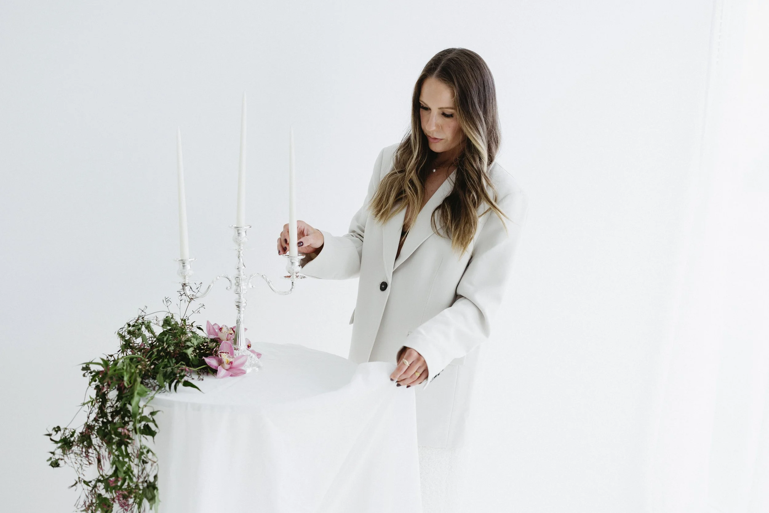 A woman in a white blazer arranging flowers and candles on a table.