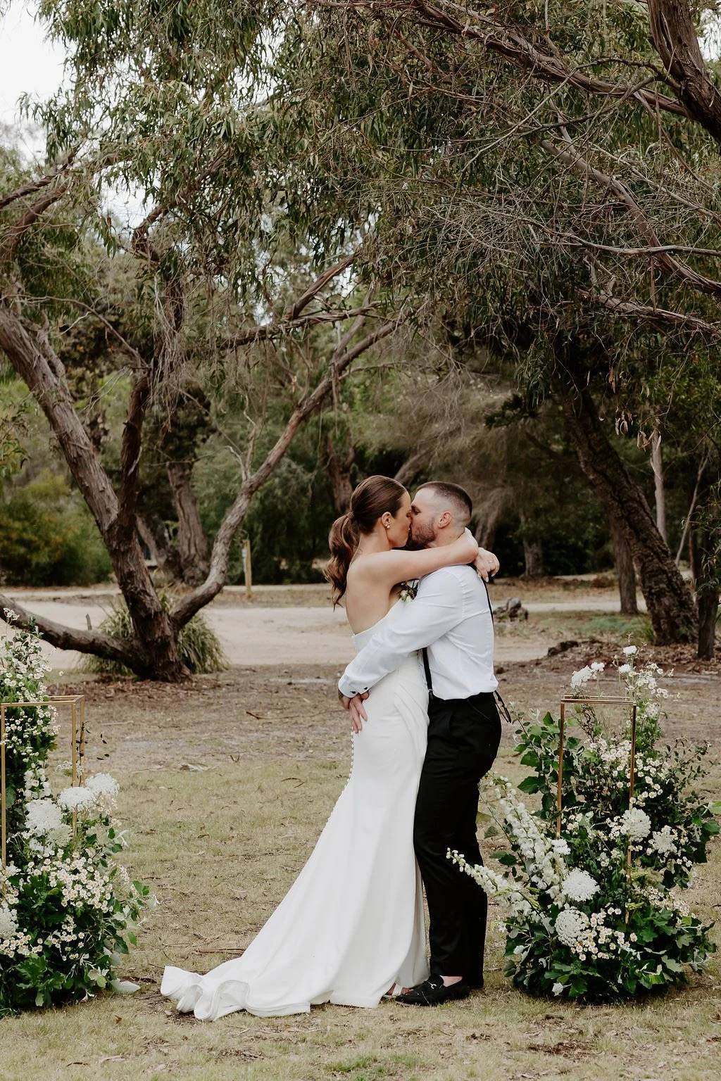 A bride and groom sharing a kiss during a wedding ceremony outdoors surrounded by trees and floral arrangements.