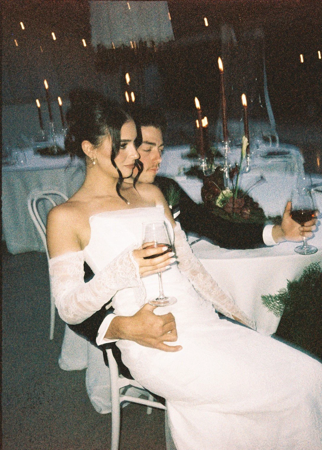 A newlywed couple in wedding attire sitting at a table, holding glasses of red wine, with lit candles and floral arrangements on the table.
