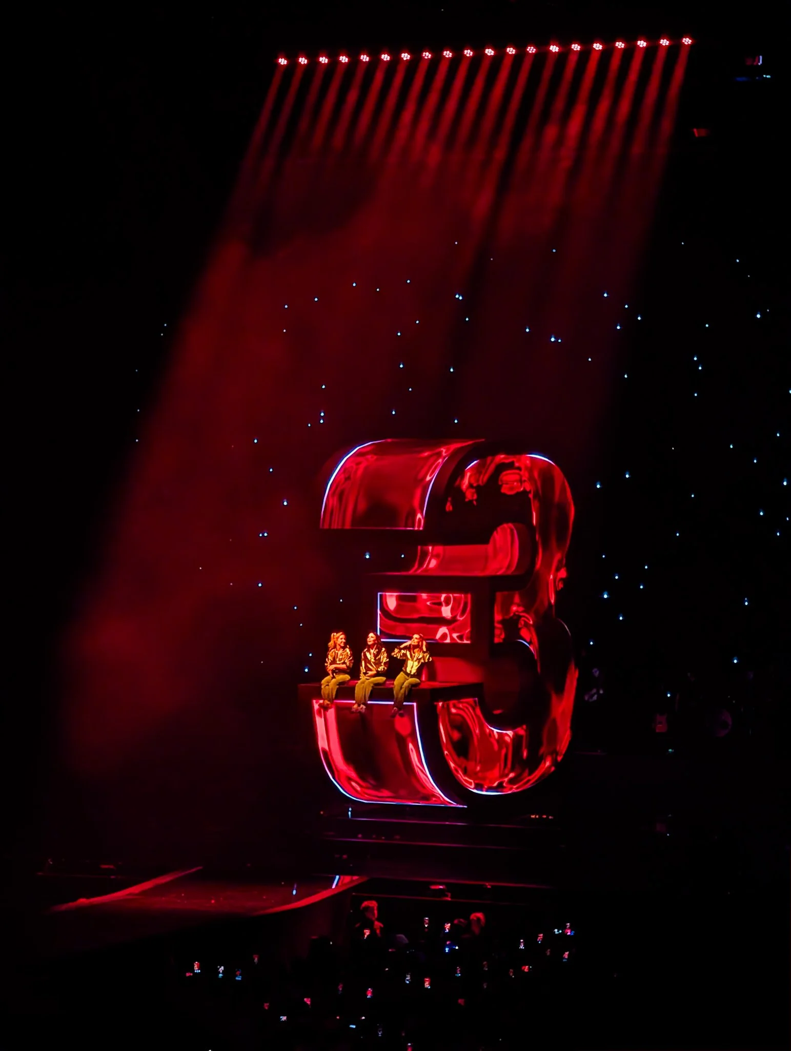 Een zwart podium met rode neon verlichte cijfers '3' en een groep mensen zittend op de cijfers tijdens een voorstelling, met een achtergrond van sterren of lichtpuntjes.