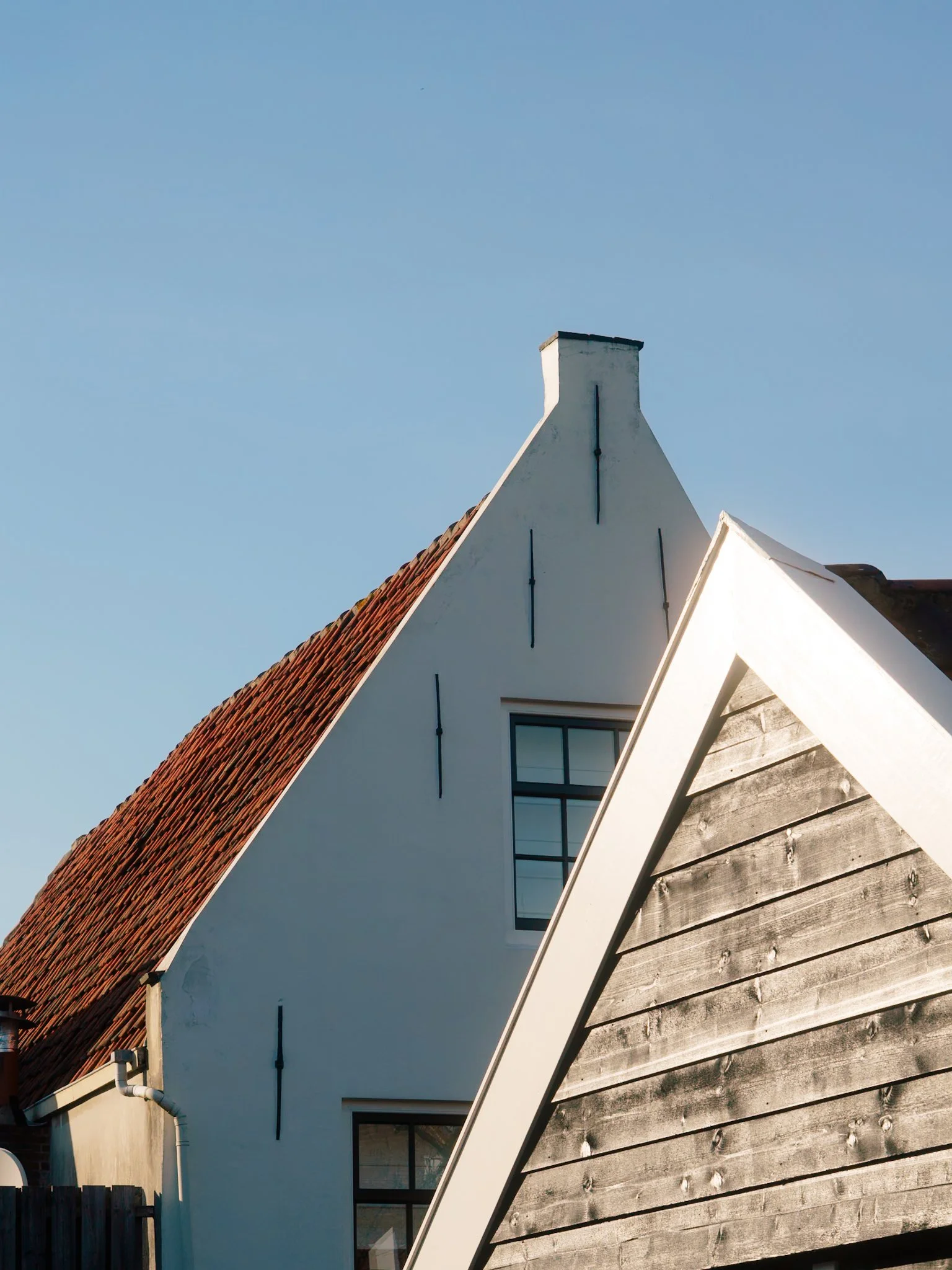 Delen van daken van huizen met verschillende materialen en kleuren, waaronder rood tegelwerk, witte muren en houten gevelbekleding, onder een blauwe hemel.