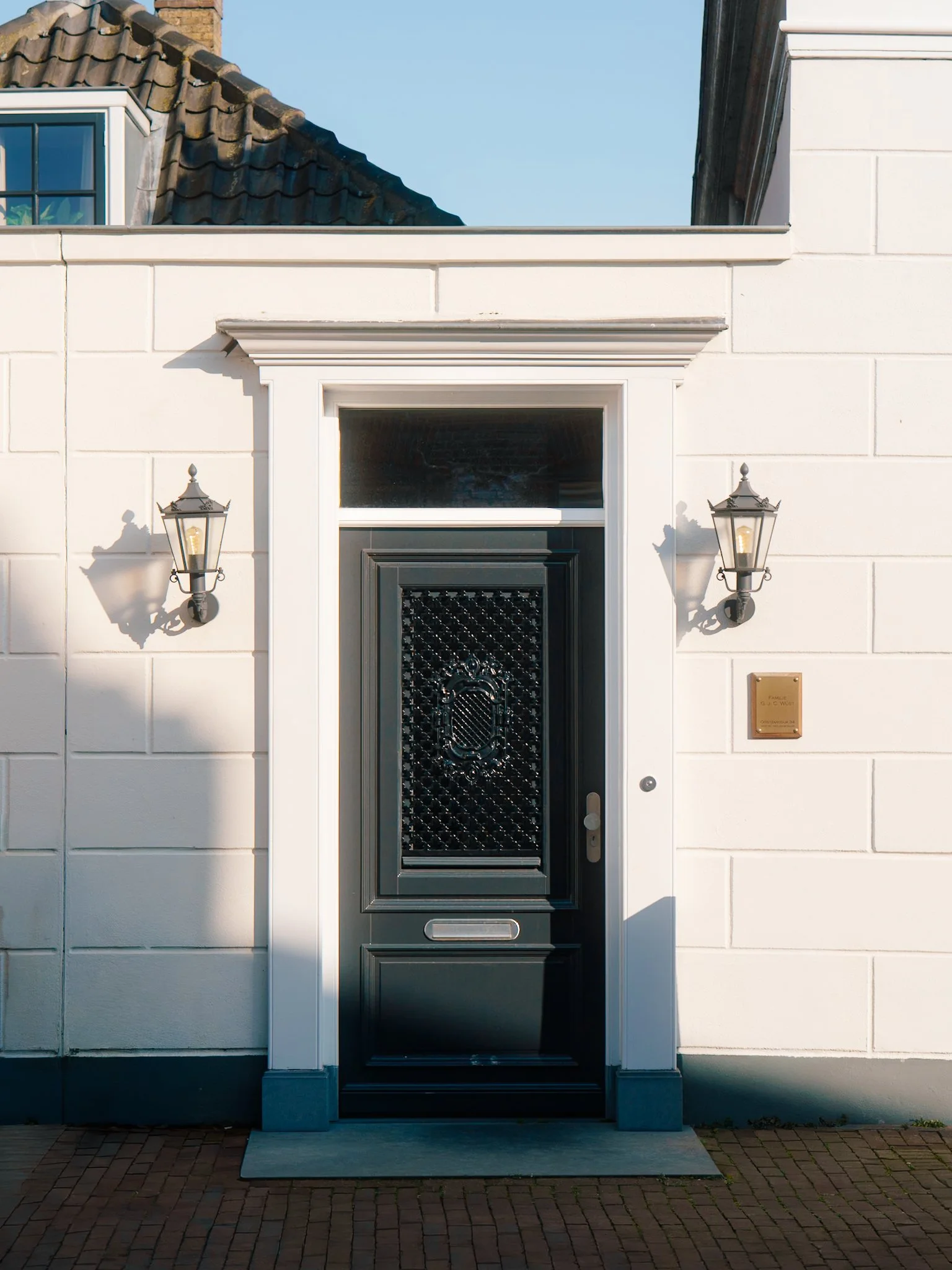 De afbeelding toont een moderne zwarte deur met een decoratief patroon, omgeven door wit gevelstenen. Aan weerszijden van de deur hangen twee zwarte lantaarnlichten. Boven de deur zit een brede raam met een witte omlijsting. Links en rechts van de de