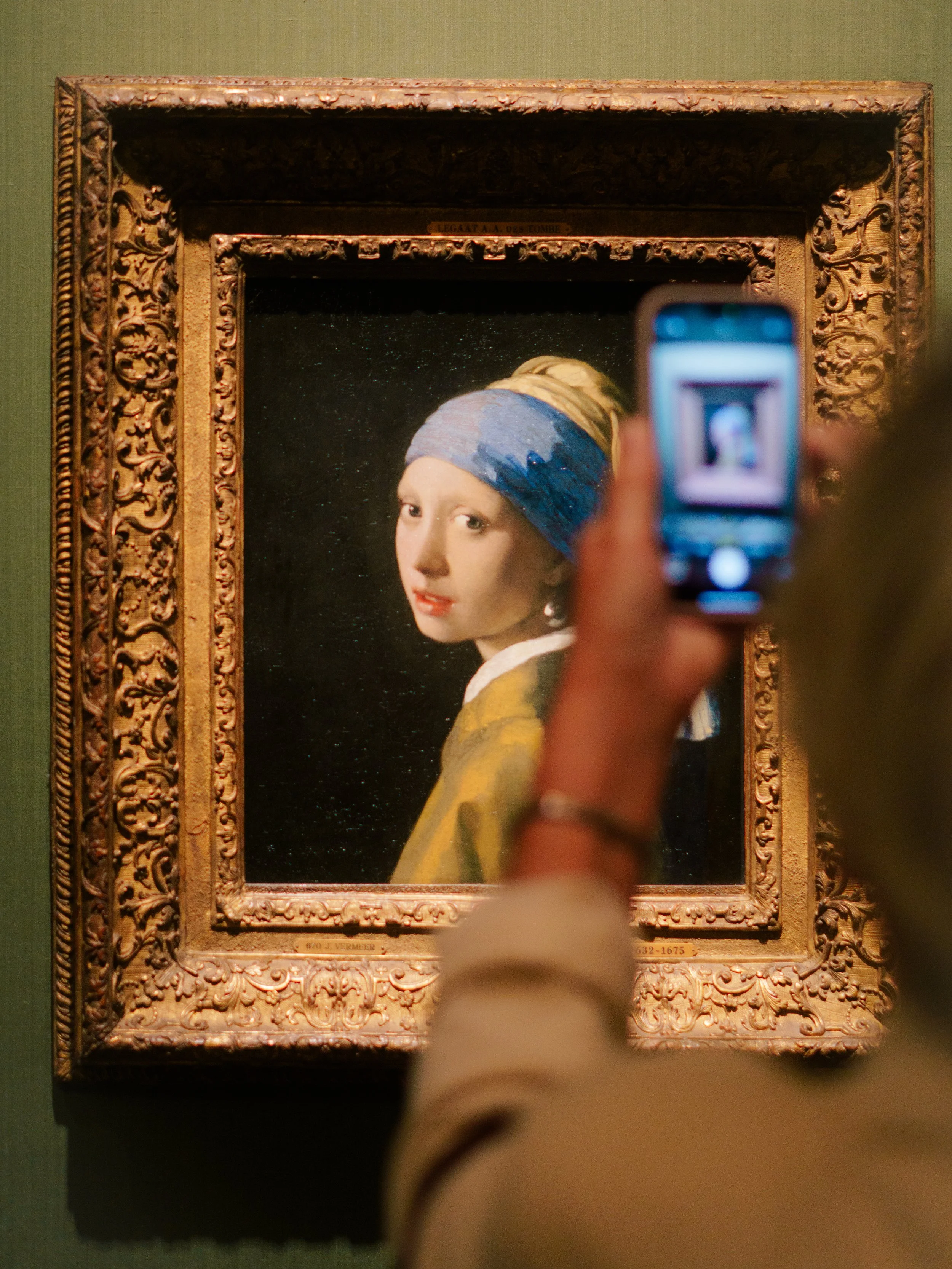 A person taking a photo of a famous portrait painting with a smartphone. The painting features a woman wearing a blue headscarf and a yellowish garment with a decorative frame.