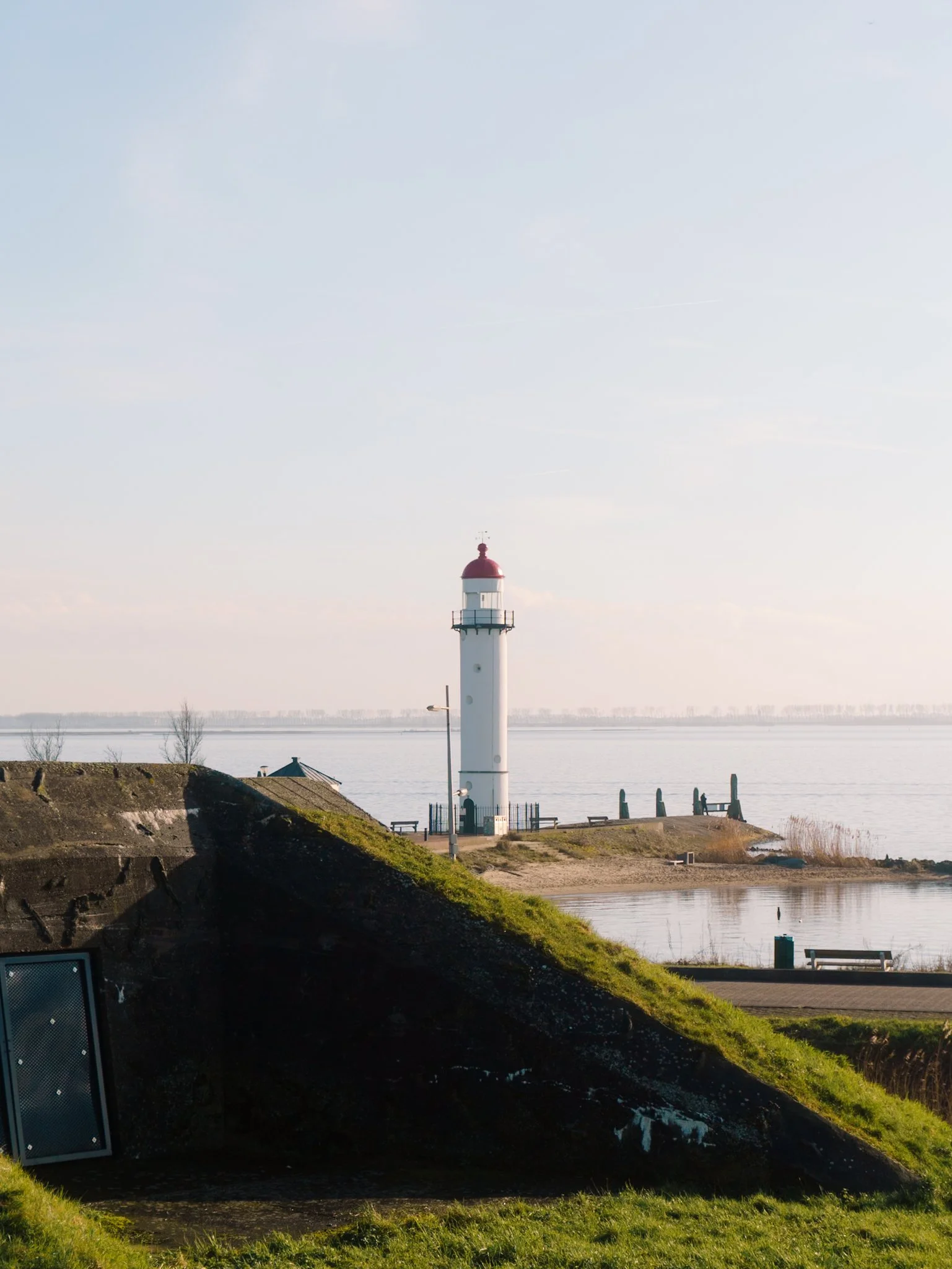 Een witte vuurtoren met een rode top staat langs de kust tijdens helder weer. In de achtergrond is water en een horizon te zien, met enkele mensen die wandelen.