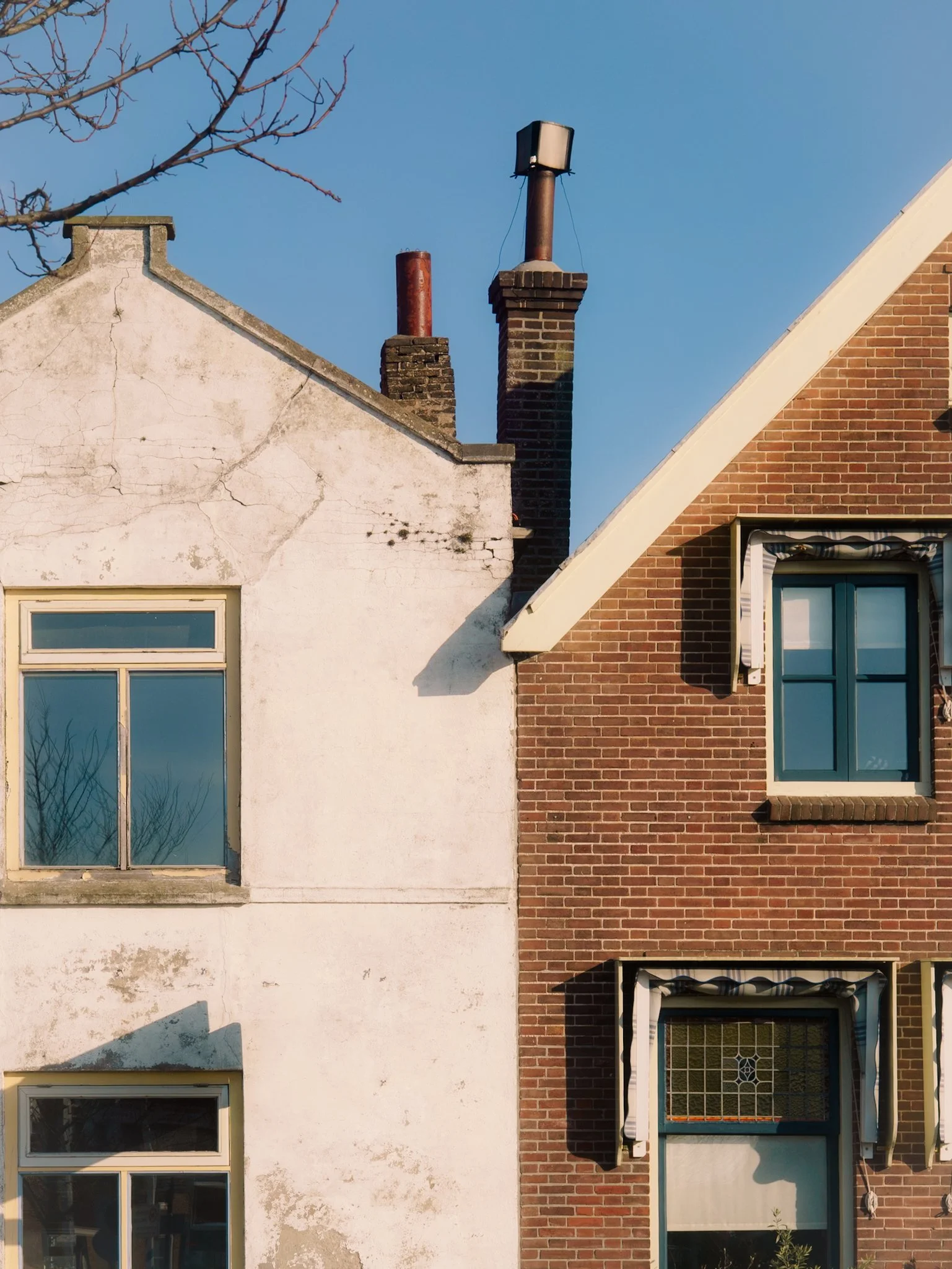 Twee huizen met verschillende gevels en daken, een met een witte muur en een ander met een bakstenen muur, onder een blauwe lucht.