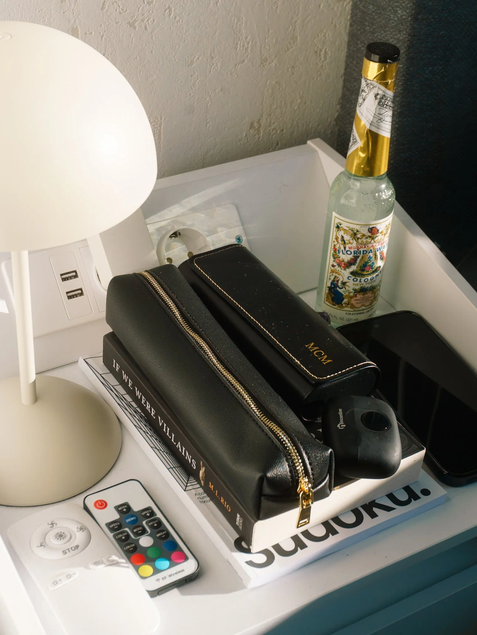 Een witte tafel met een witte lamp, afstandsbediening met meerdere gekleurde knoppen, een zwart make-up toilettasje met gouden ritssluiting, een boek met de titel 'Stadstaten', een mobiele telefoon, een lege flessenwasser en een bouquet van bloemen o
