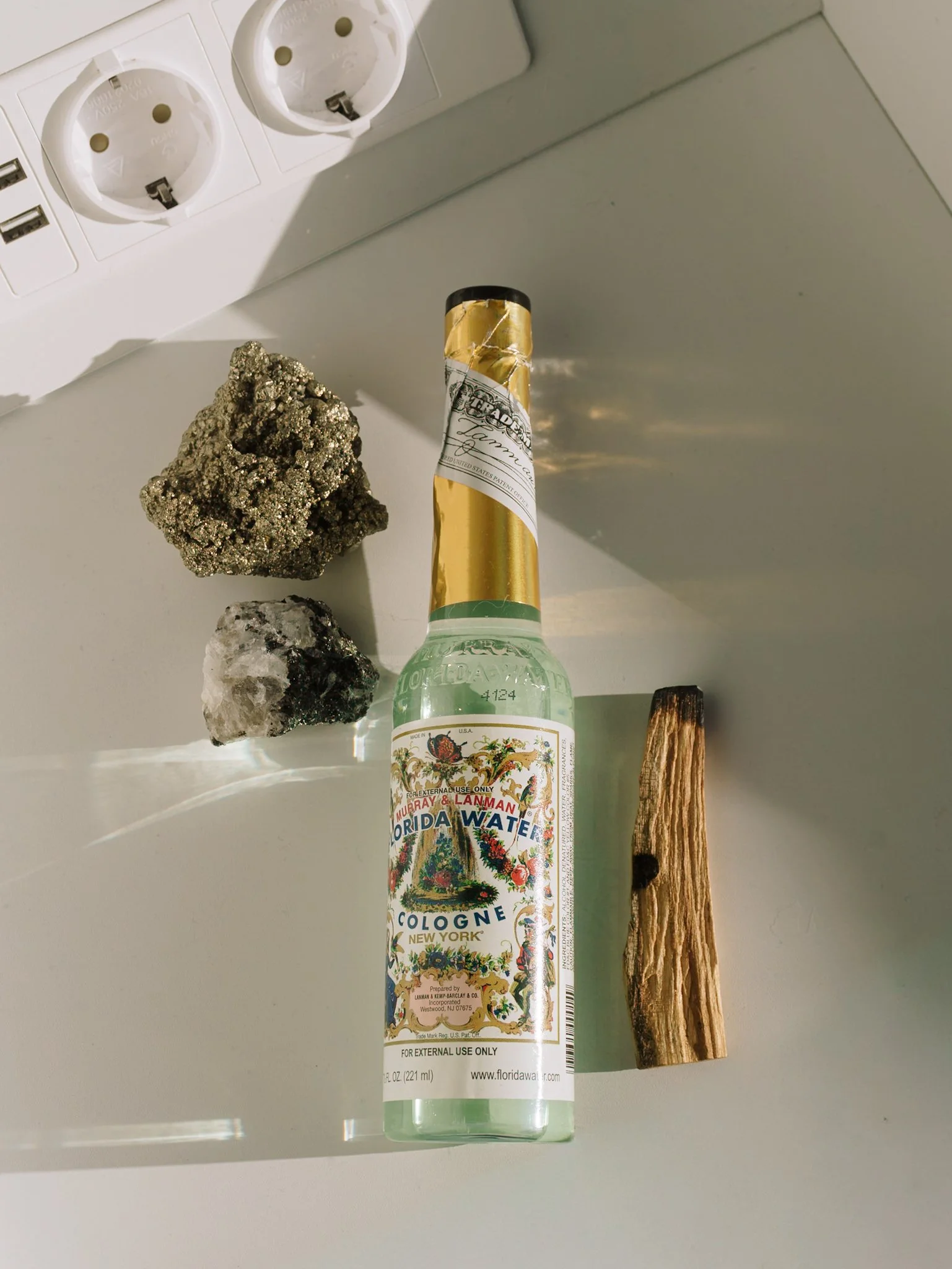 Een flesje Florida Water cologne, twee mineralenstenen en een stuk hout op een wit oppervlak. Een elektrische stopcontact met twee stopcontacten en twee USB-poorten is op de achtergrond zichtbaar.