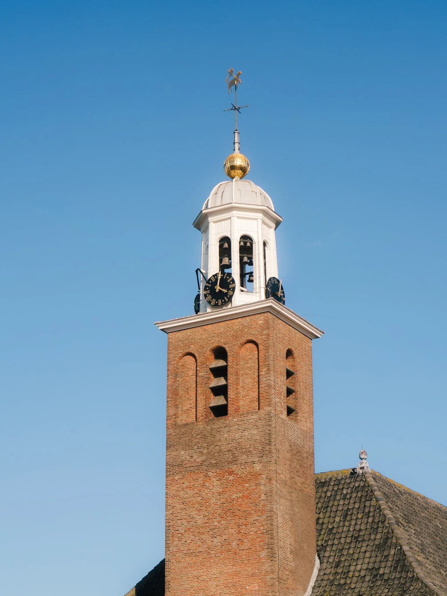 Een toren met een klok en een weerhaan op de top, tegen een heldere blauwe lucht.
