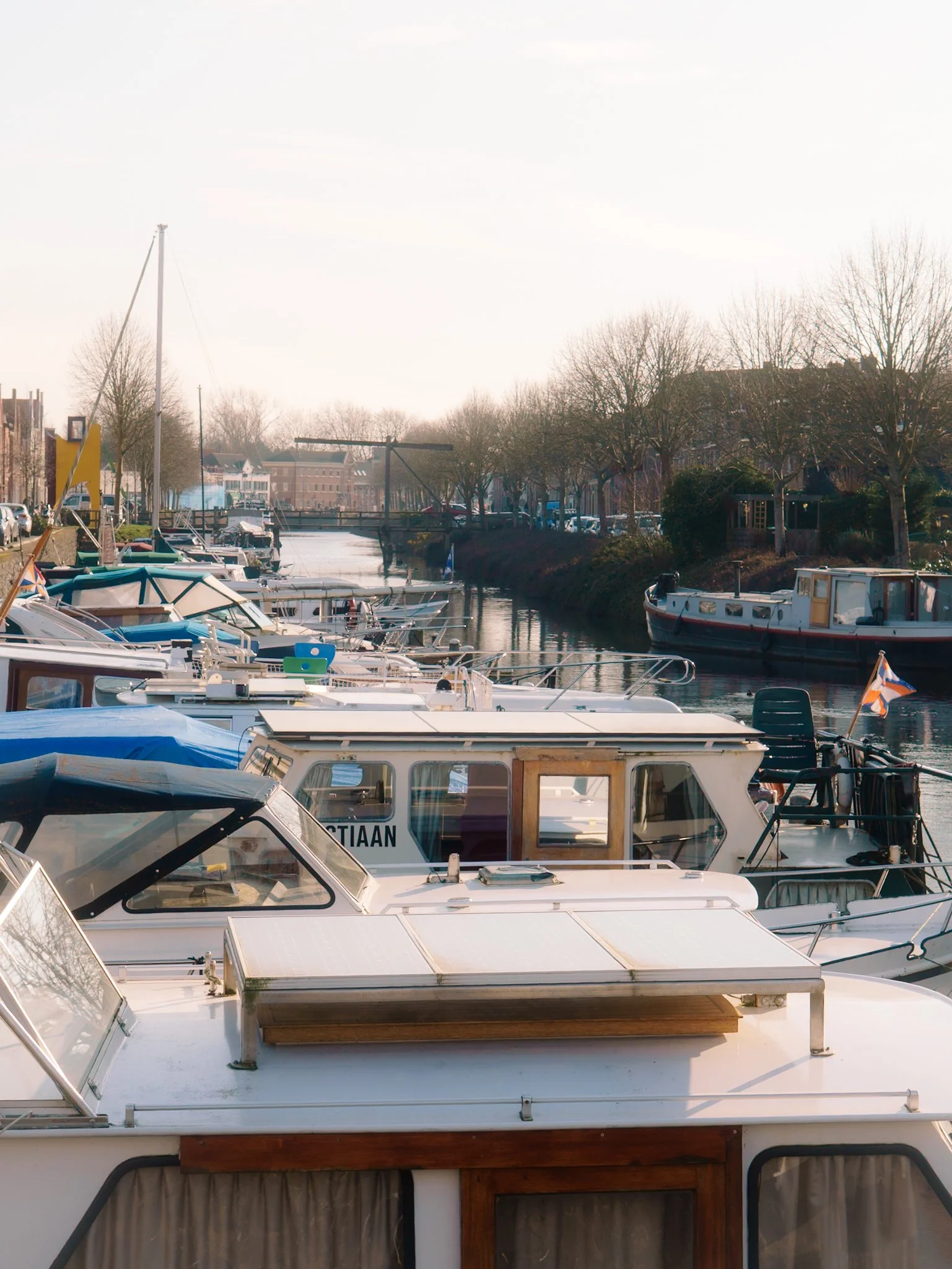 Boots en huizen langs een kanaal met bomen en een brug, onder een heldere hemel.
