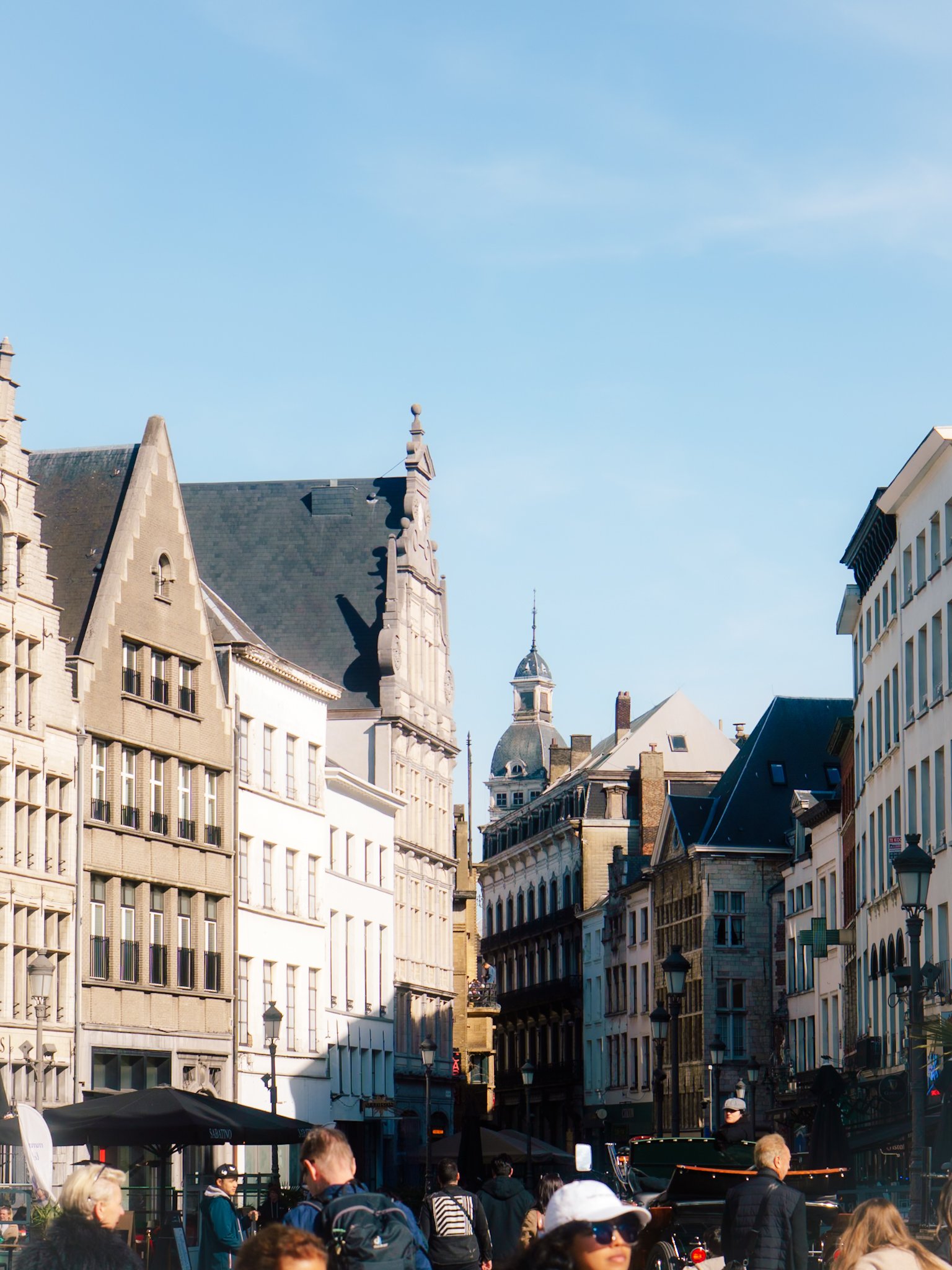 Een drukke straat in een stad met historische gebouwen en mensen die wandelen. De lucht is helder blauw en er zijn terrassen met parasols.