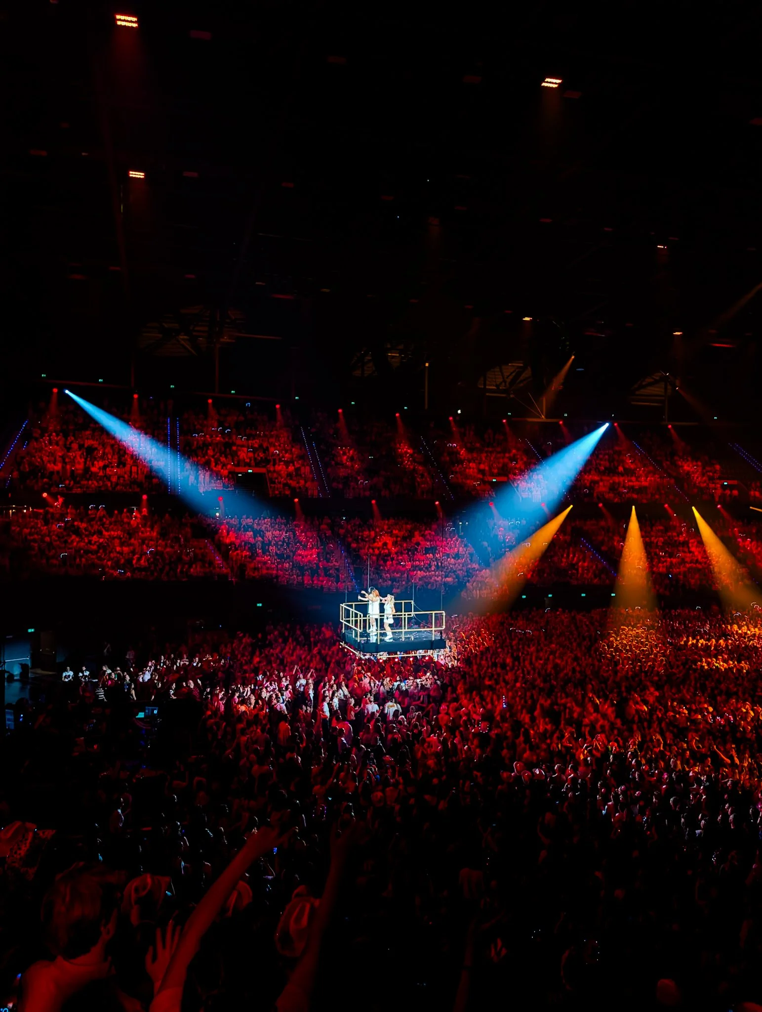 Een groot binnenarena met een publiek en een toneel met twee performers op een hijsplatform, omringd door kleurrijke podiumverlichting.