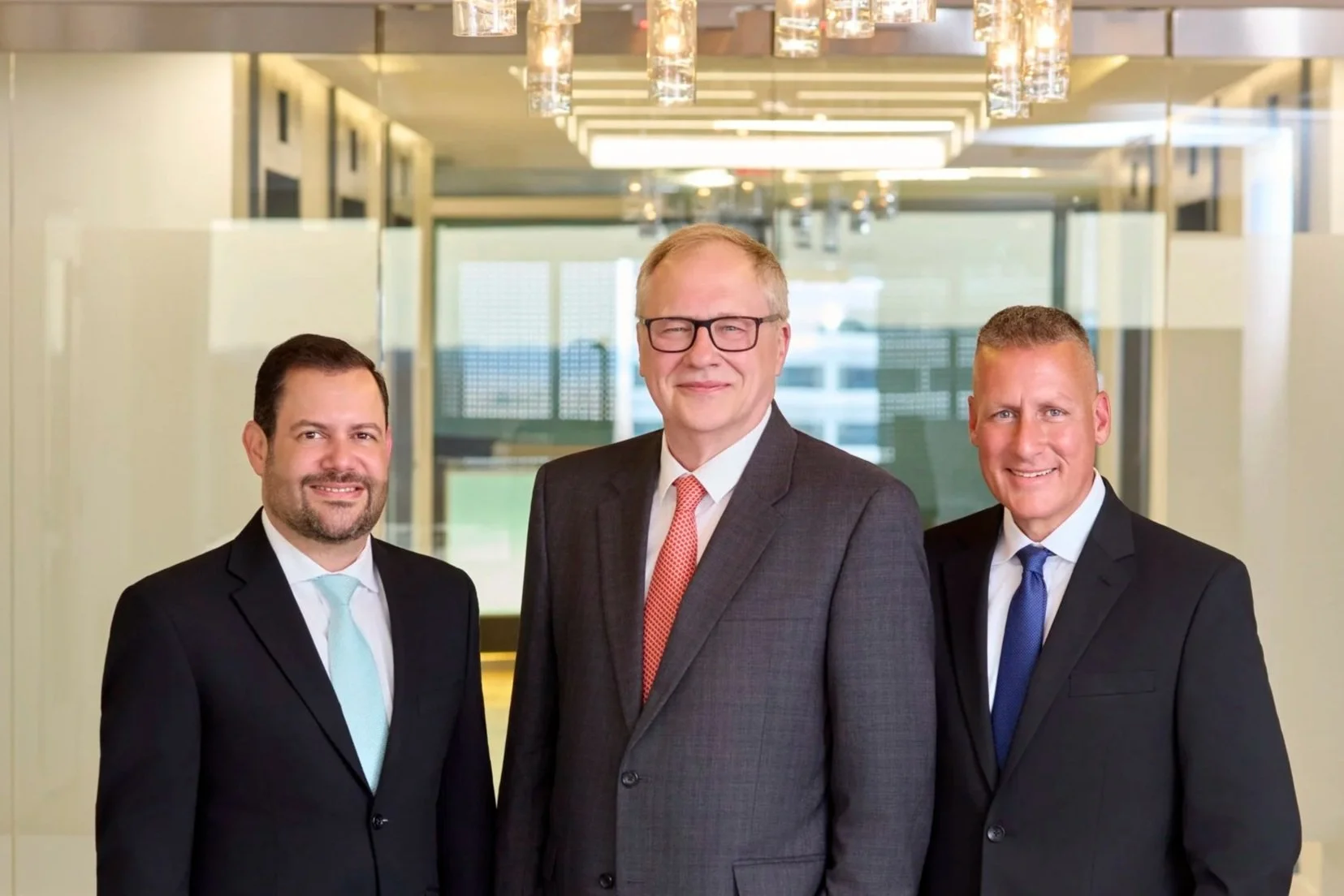 Three men in business suits standing in an office corridor, smiling at the camera.