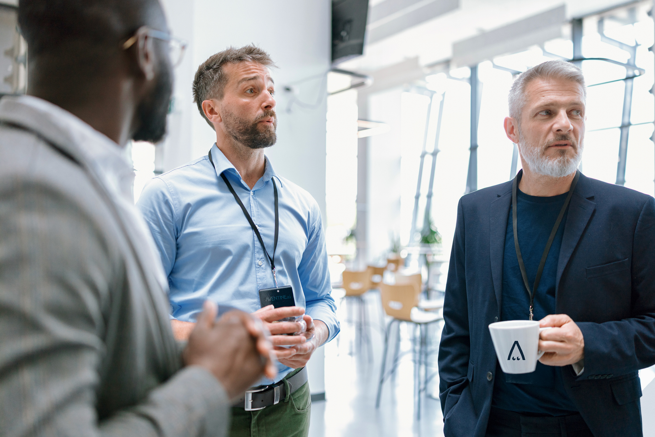 Three men are engaged in conversation during a business meeting or networking event in a modern, well-lit space. One man is holding a coffee mug with a logo, and another is holding a water bottle. All are wearing business casual or professional attire with name tags or lanyards. The background features large windows, allowing natural light to illuminate the scene.