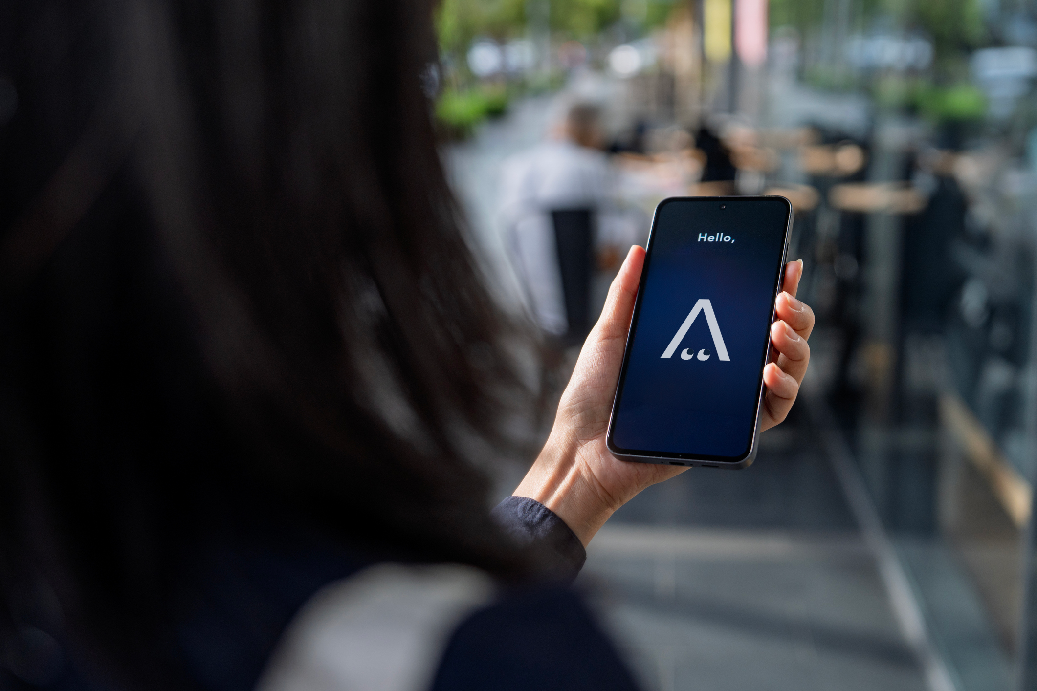Person holding a smartphone with a dark screen displaying the word 'Hello,' and a stylized logo with a triangular shape and dots, in a busy indoor setting.