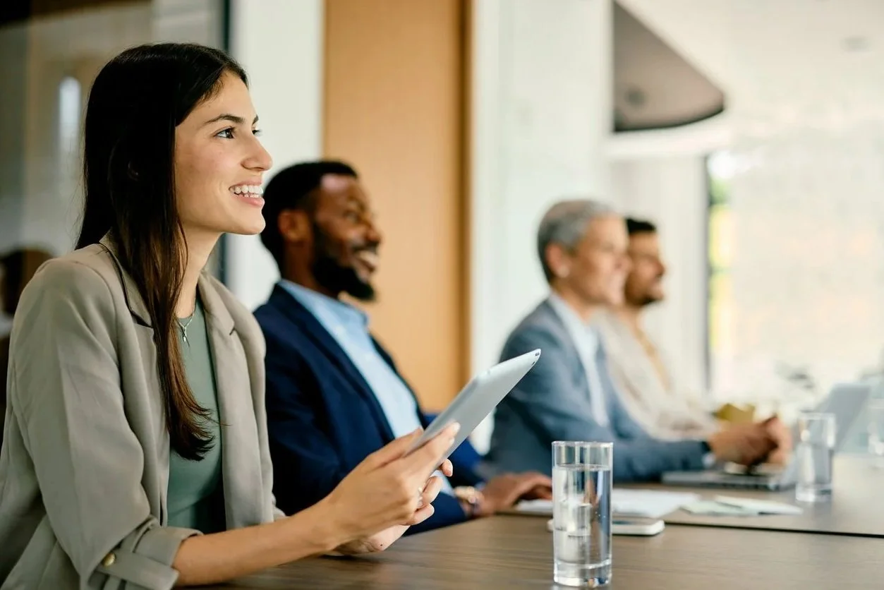 A diverse group of four professionals sitting at a conference table during a meeting, with the woman in the foreground smiling and holding a tablet.