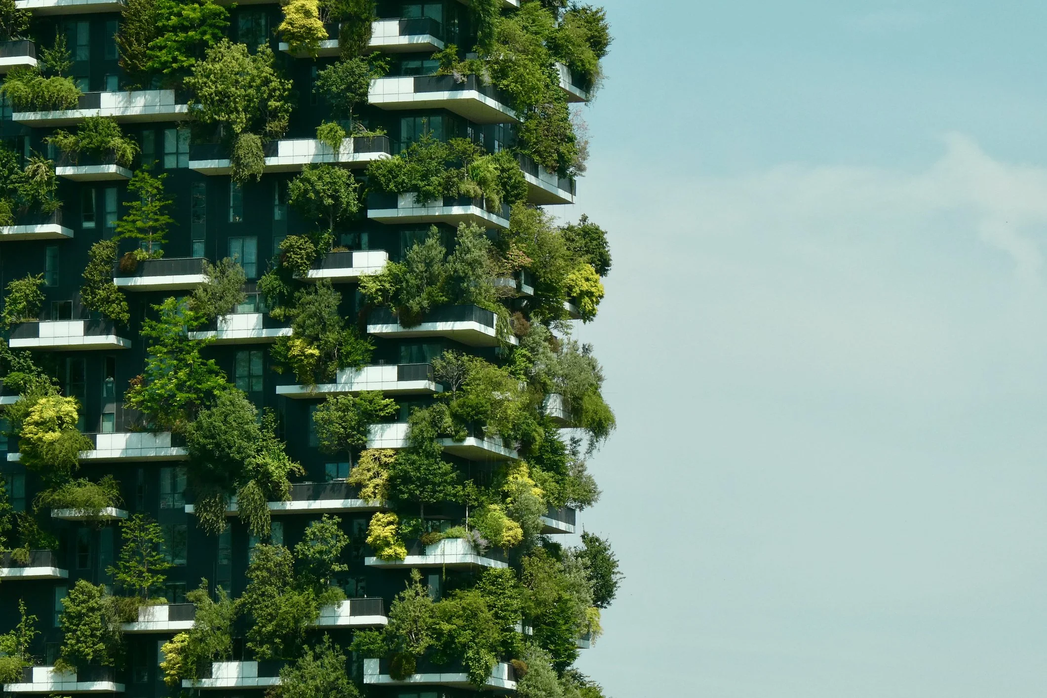 A modern high-rise building with numerous balconies covered in lush green trees and plants, set against a light blue sky.